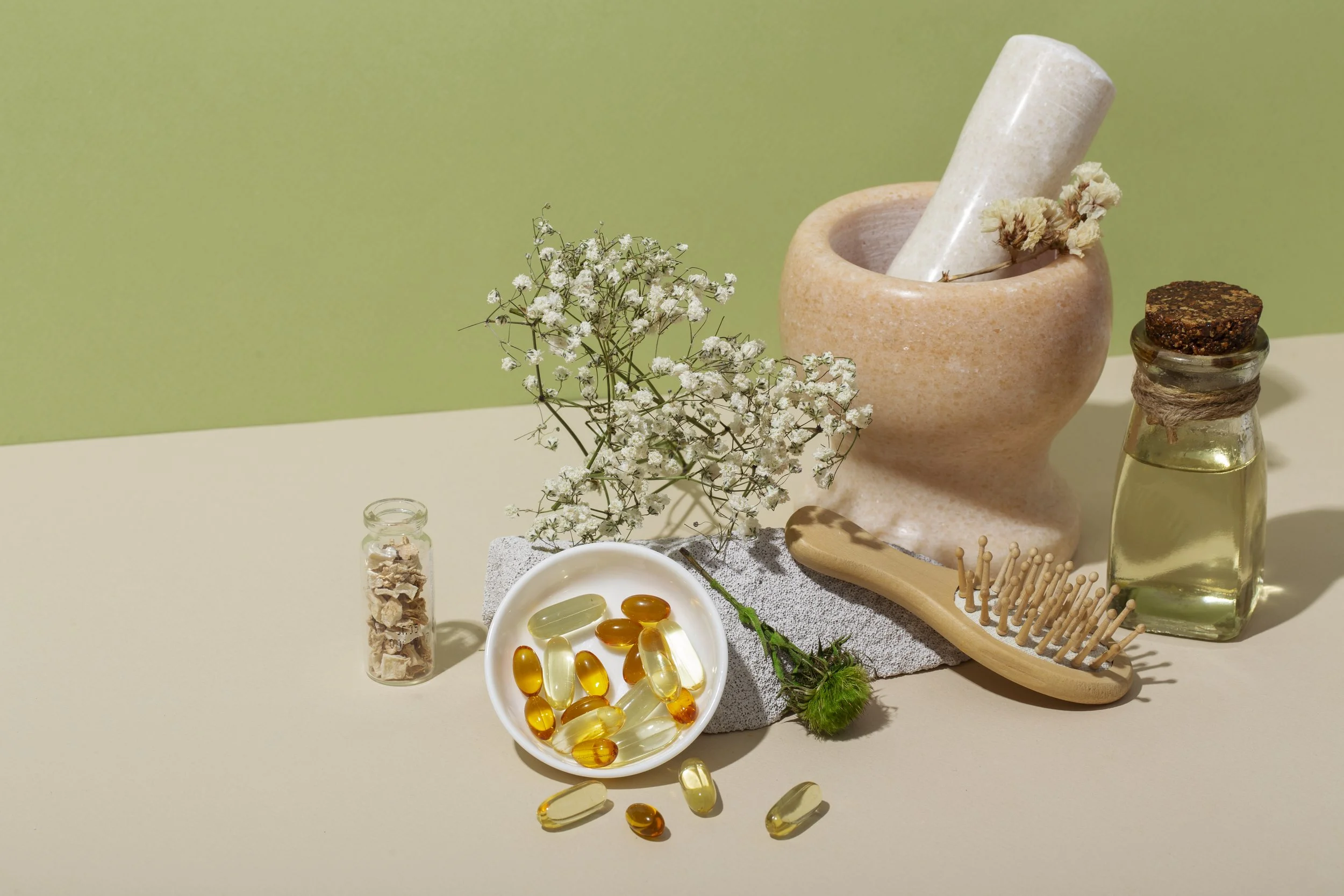 Arrangement of natural remedies, supplements, and wellness items including a small jar of dried herbs, a bowl of gel capsules, a sprig of white flowers, a wooden brush, a pink mortar and pestle with a flower, and small glass bottles with oils or tinctures, on a white surface with a green background.