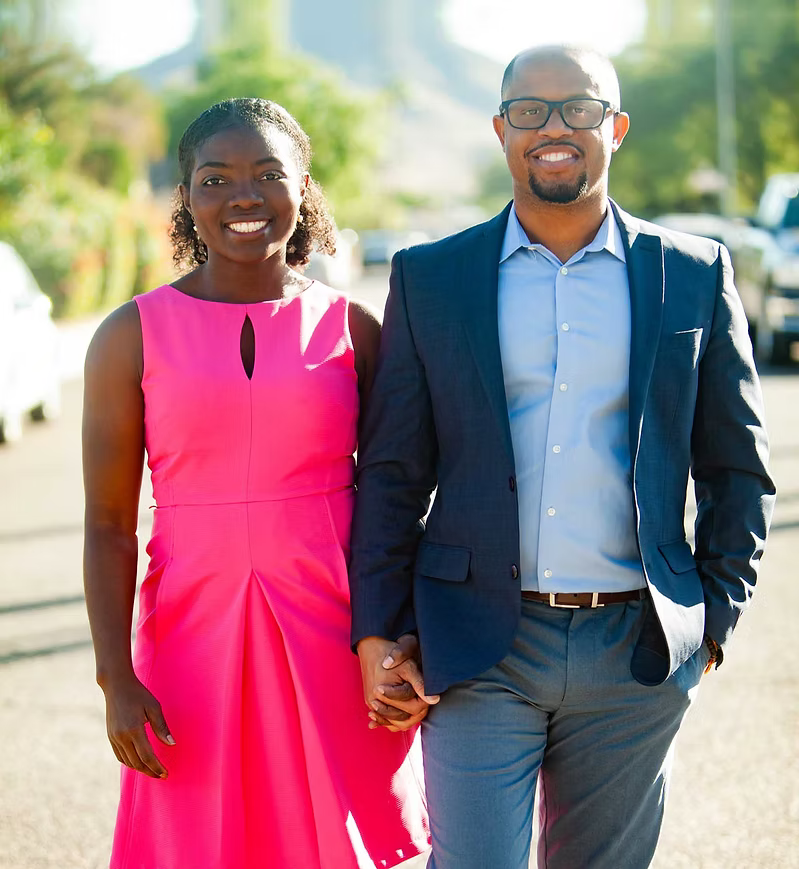 Danielle and Franz Delima of Arise & Own holding hands outdoors with trees and cars in the background. The woman wears a pink dress, and the man wears a navy blue suit with glasses.