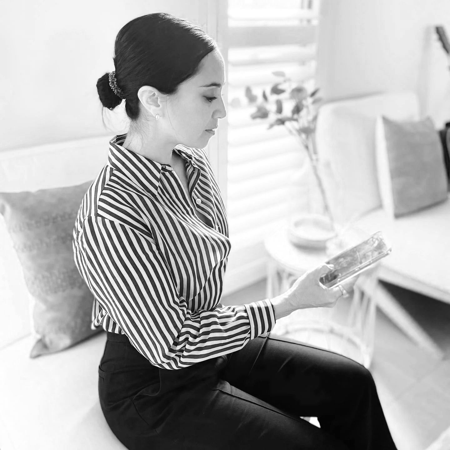 Black and white photo of a woman sitting on the floor, looking at her smartphone, in a room with a sofa, decorative pillows, a side table with a flower vase, and window blinds.