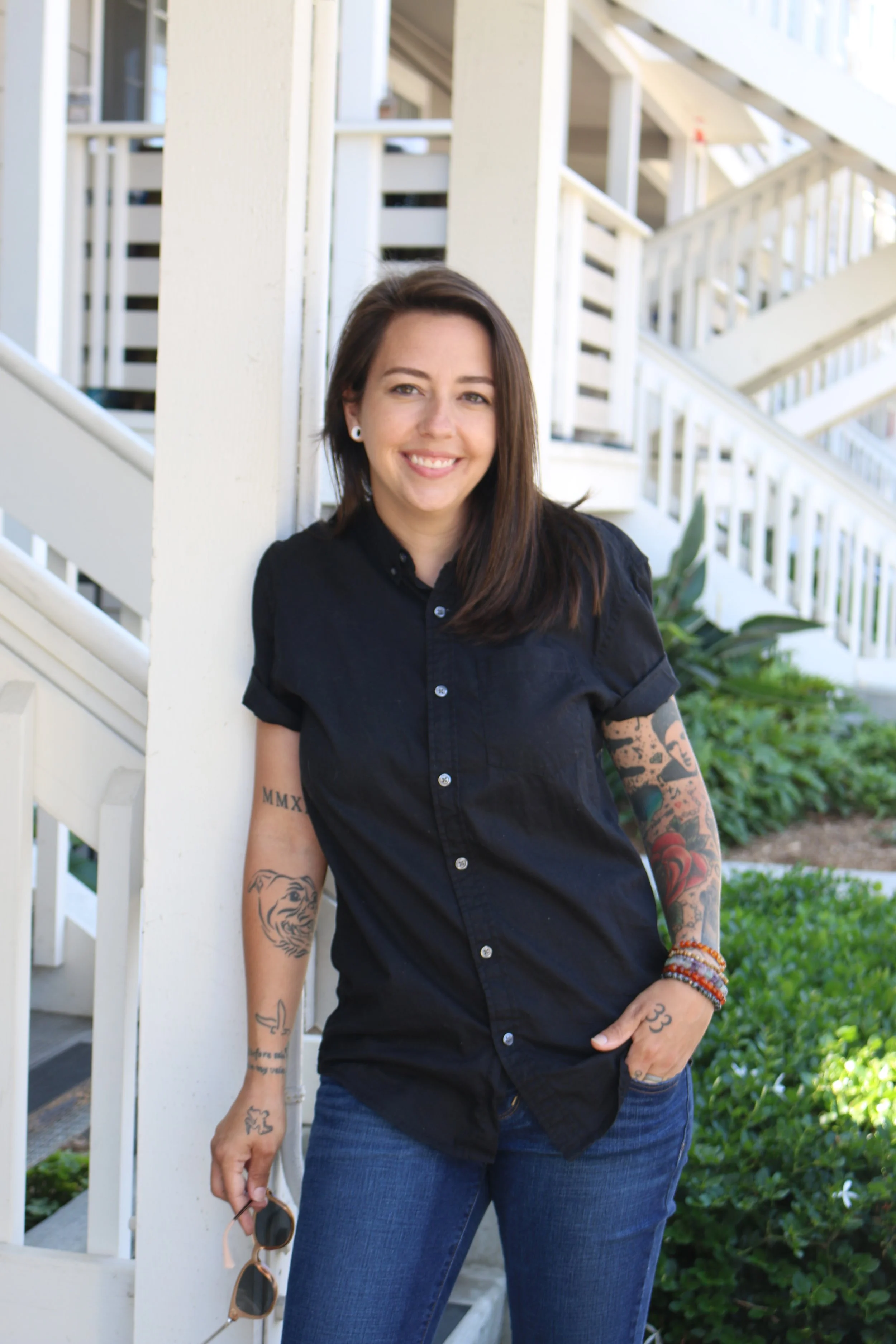 A woman with brown hair, tattoos on both arms, wearing a black button-up shirt and blue jeans, standing outdoors near a white stairway and greenery, smiling at the camera.