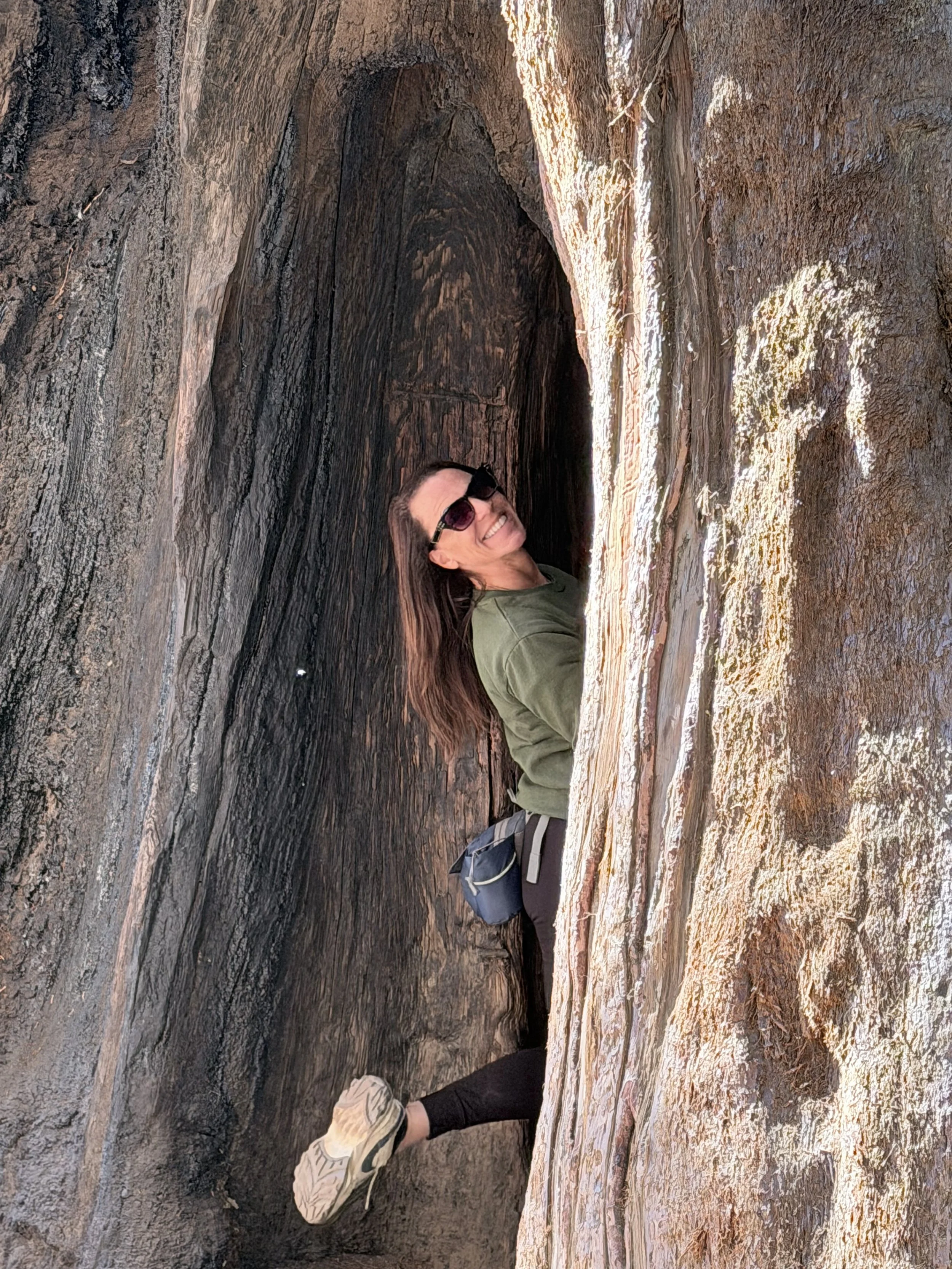 Smiling woman with sunglasses, green jacket, and backpack sitting in a narrow, deep rock crevice.