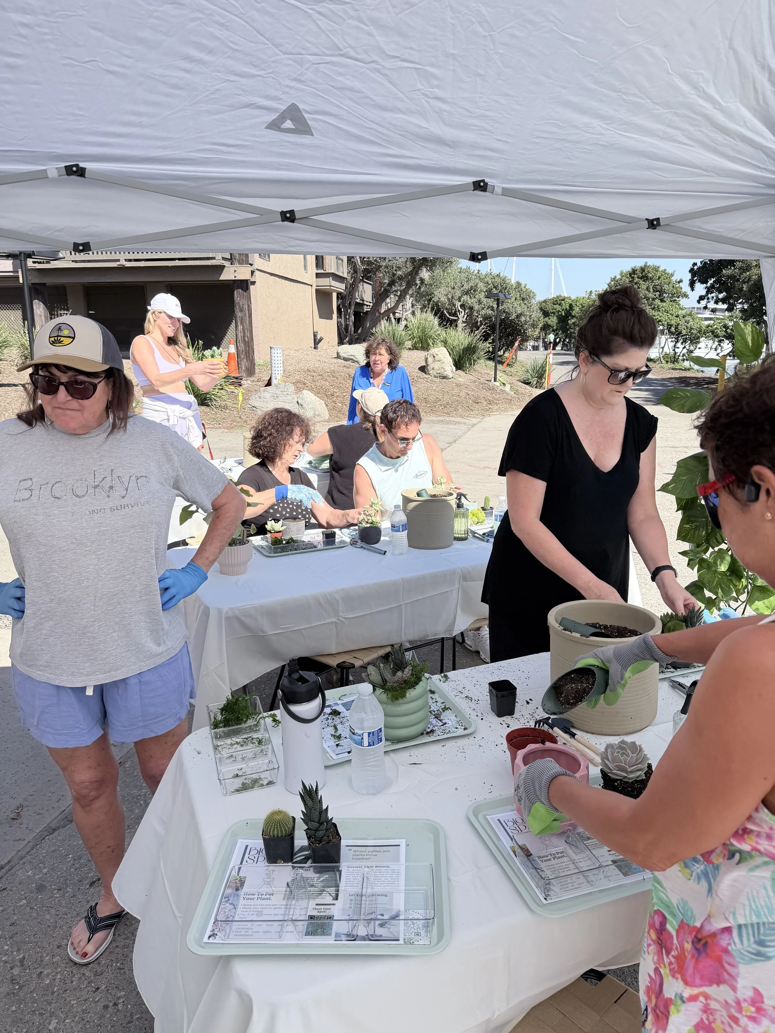 Group of people participating in a gardening or plant workshop outdoors under a tent, with tables, plants, and gardening tools.