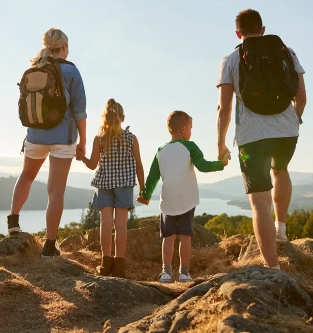 A family of five, including two adults and three children, hiking on a rocky trail overlooking a lake and mountains during sunset.