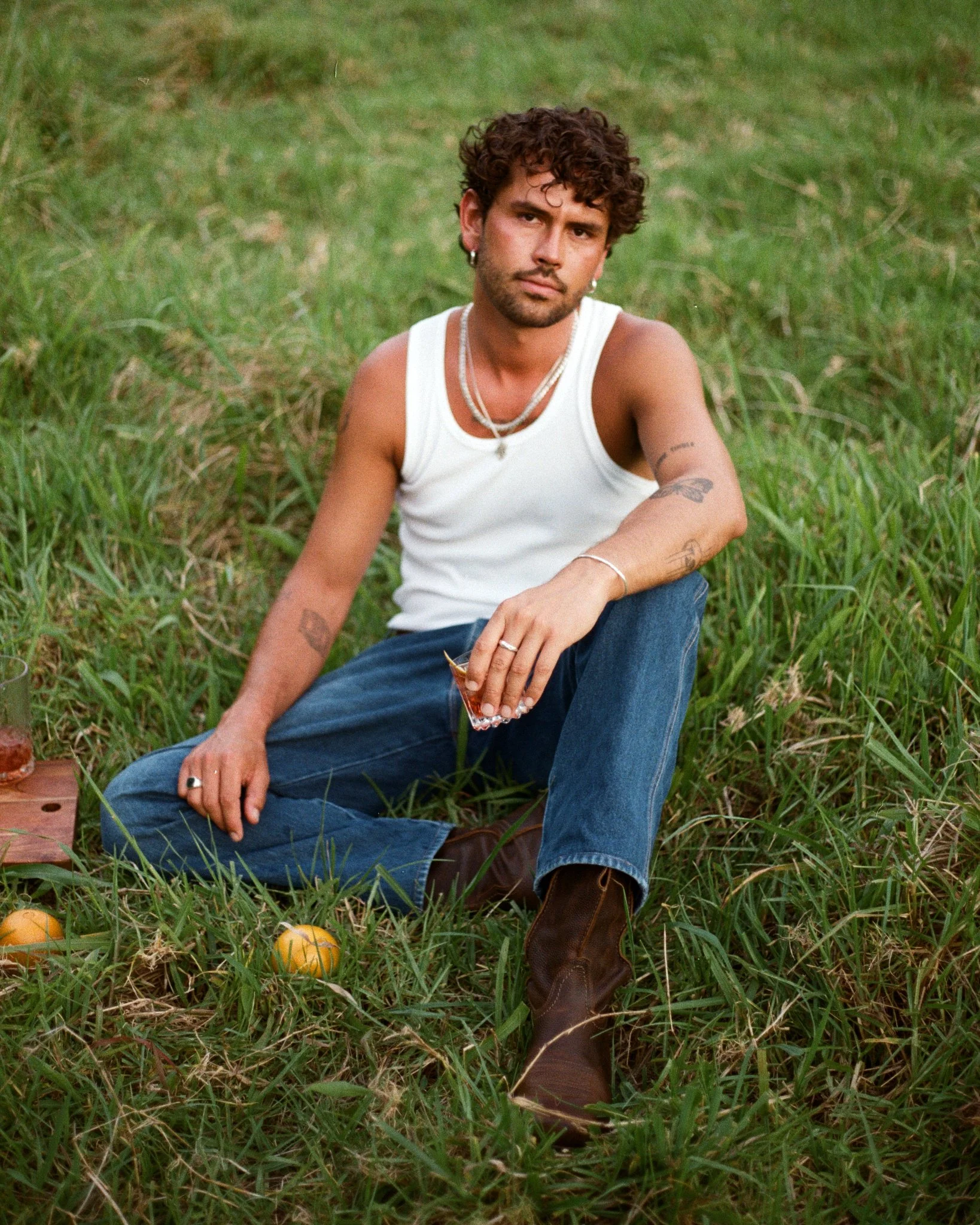 A man with curly hair, wearing a white tank top, blue jeans, and brown boots, is sitting on the grass in a field. He has tattoos, jewelry, and is holding a glass in his hand. There are some small oranges and a wooden object nearby.