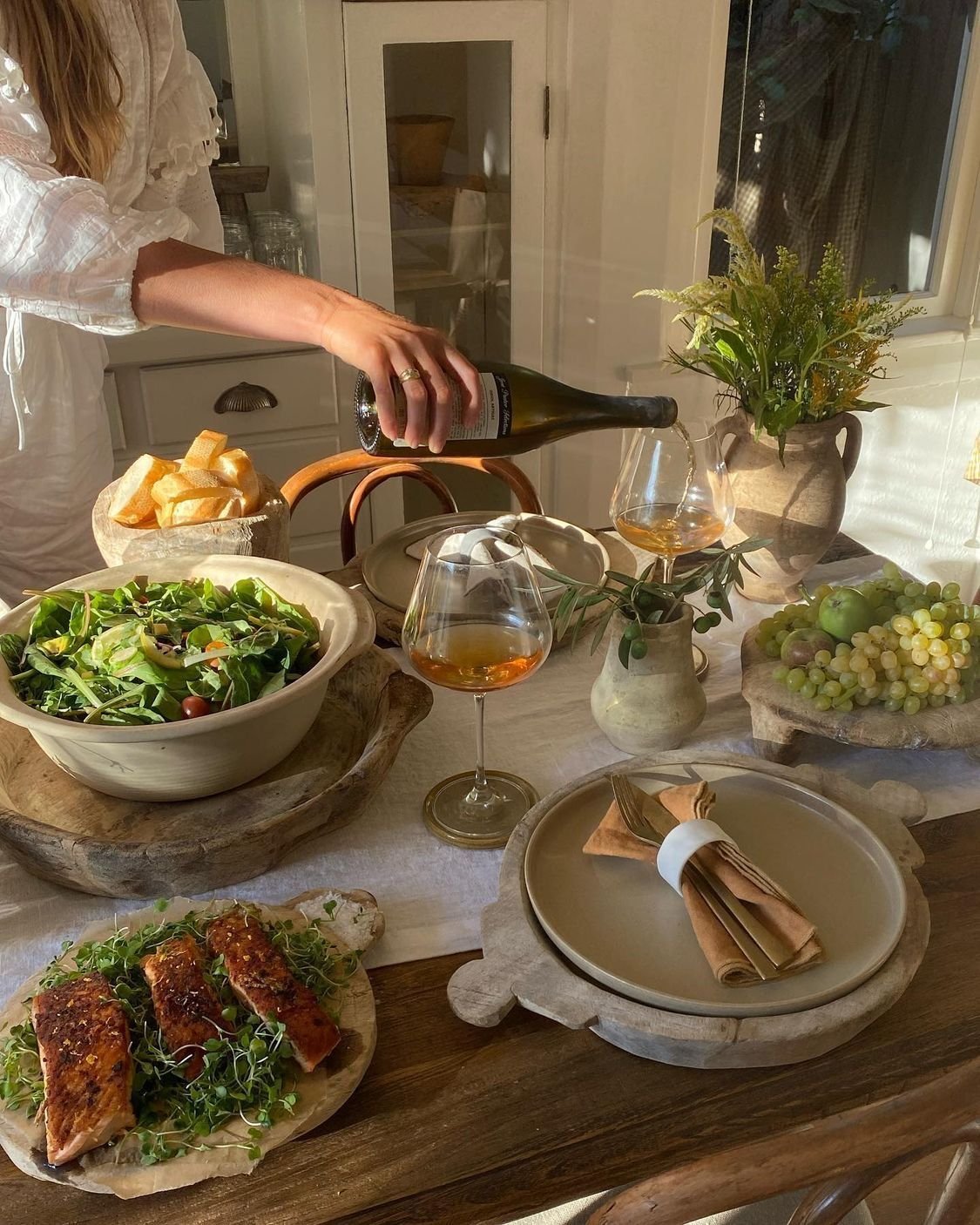 Person pouring wine at a dining table set with a salad, bread, grapes, and plates, with sunlight streaming in.