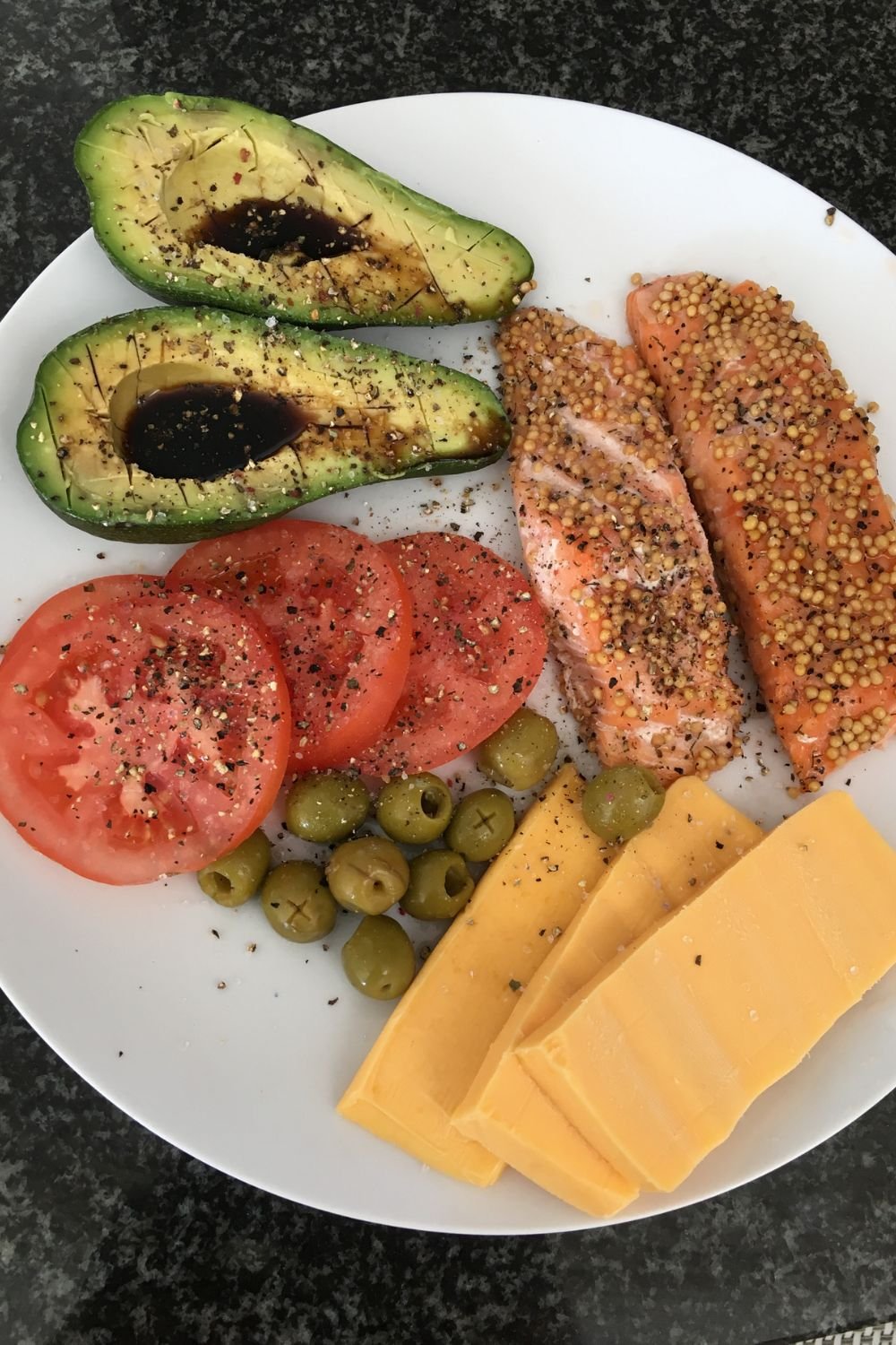 Plate with avocado halves topped with balsamic glaze, sliced tomatoes, green olives, slices of cheddar cheese, and two pieces of salmon covered with mustard seeds and black pepper
