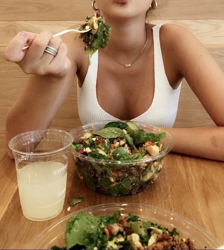 Person with a forkful of salad, sitting at a table with a salad bowl, a cup of a yellow beverage, and a lettuce wrap or sandwich in front. The person is wearing a white tank top, necklace, and earrings.