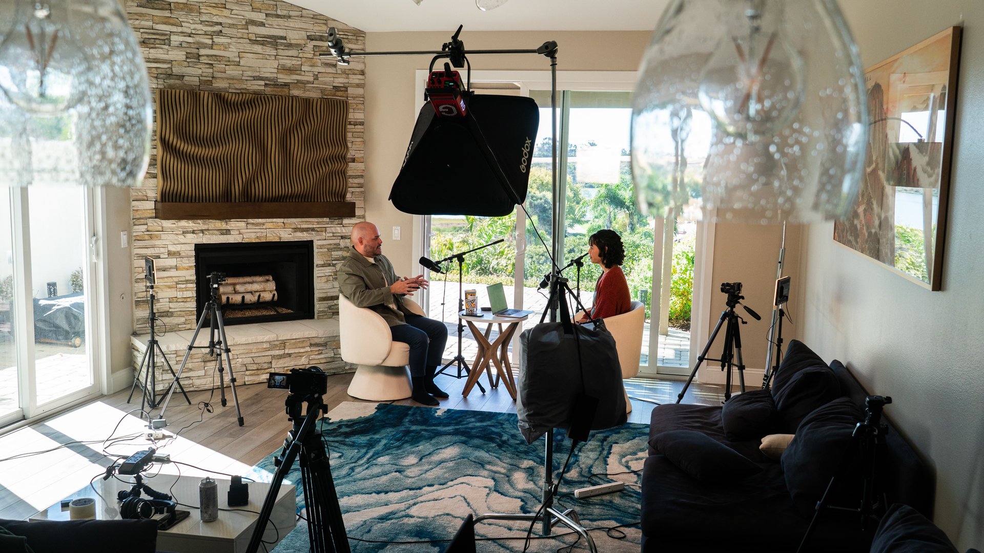 Two people are recording an interview in a living room with a stone fireplace, large windows, and a blue patterned rug. Various cameras, tripods, and lighting equipment surround them.