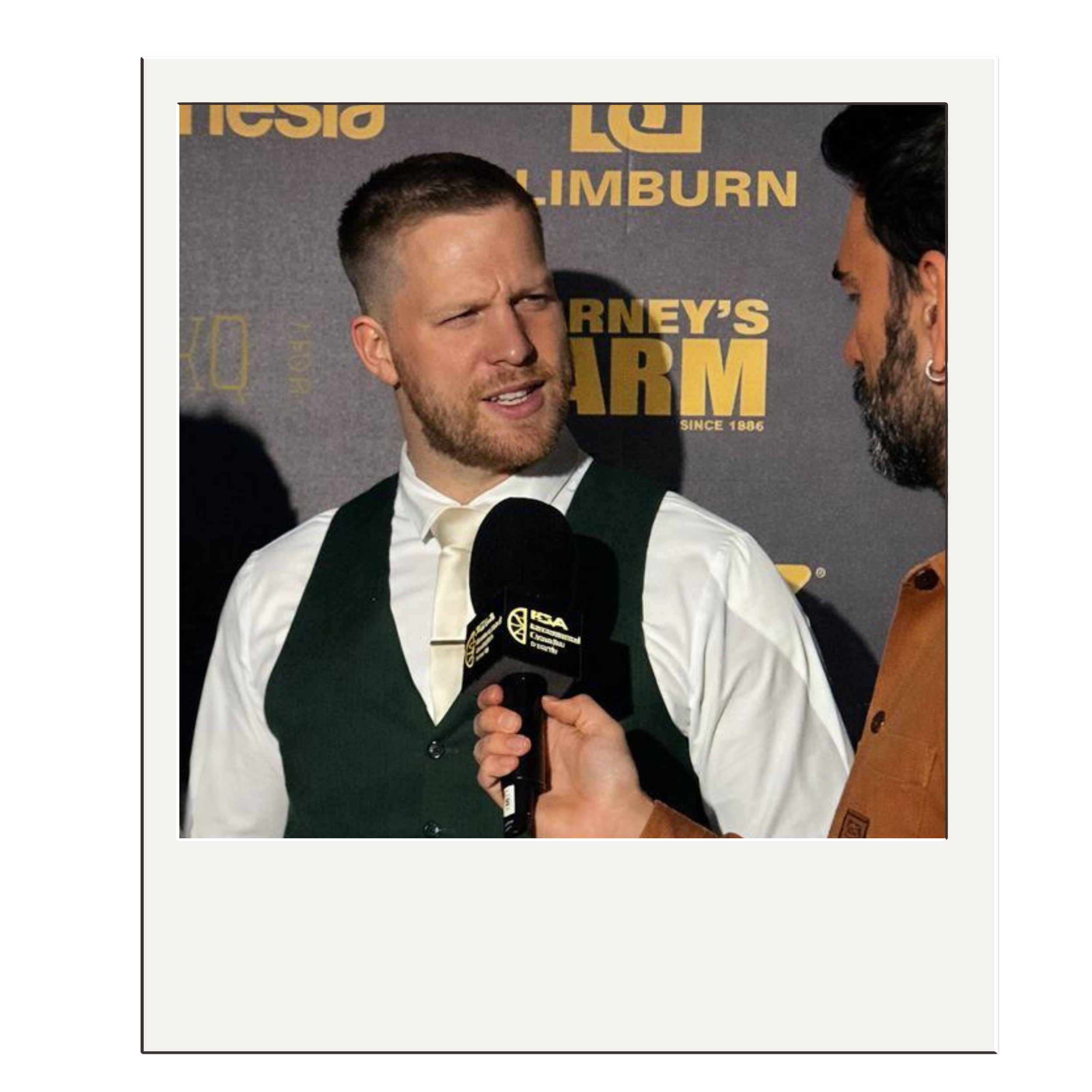 Two men having a conversation at an event. One man is being interviewed with a microphone that has the NCAA logo. The background features a sign for Barstool's Barstool Sportsbook and Theside.