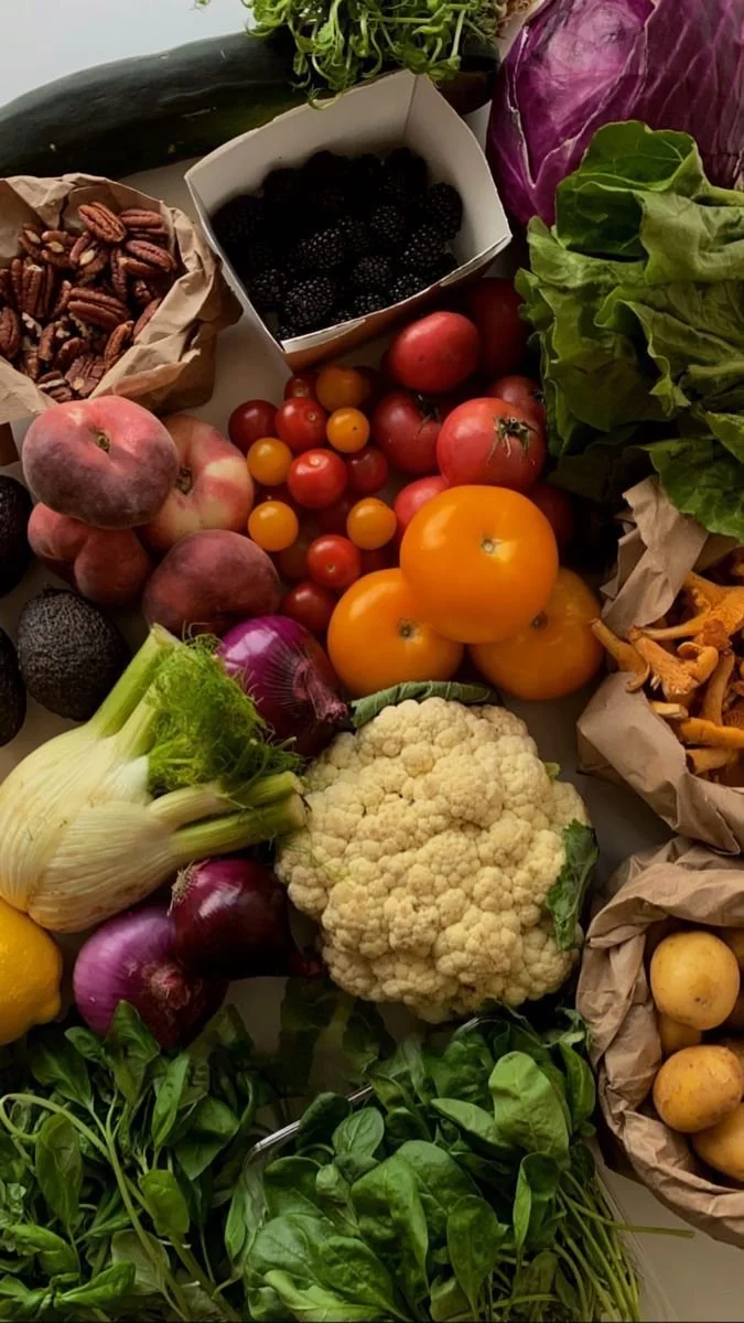 Assorted fresh vegetables and fruits including cauliflower, red onions, purple cabbage, cherry tomatoes, heirloom tomatoes, blackberries, pecans, lettuce, zucchini, and potatoes arranged on a table.