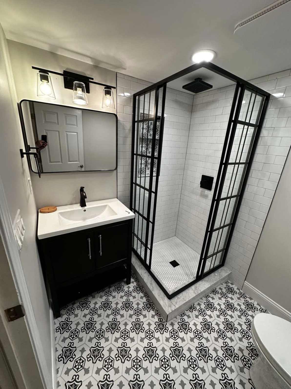 Small bathroom with patterned tile floor, black vanity with white sink, mirror above, modern lighting, glass shower with black frame, white subway tile walls, and toilet.