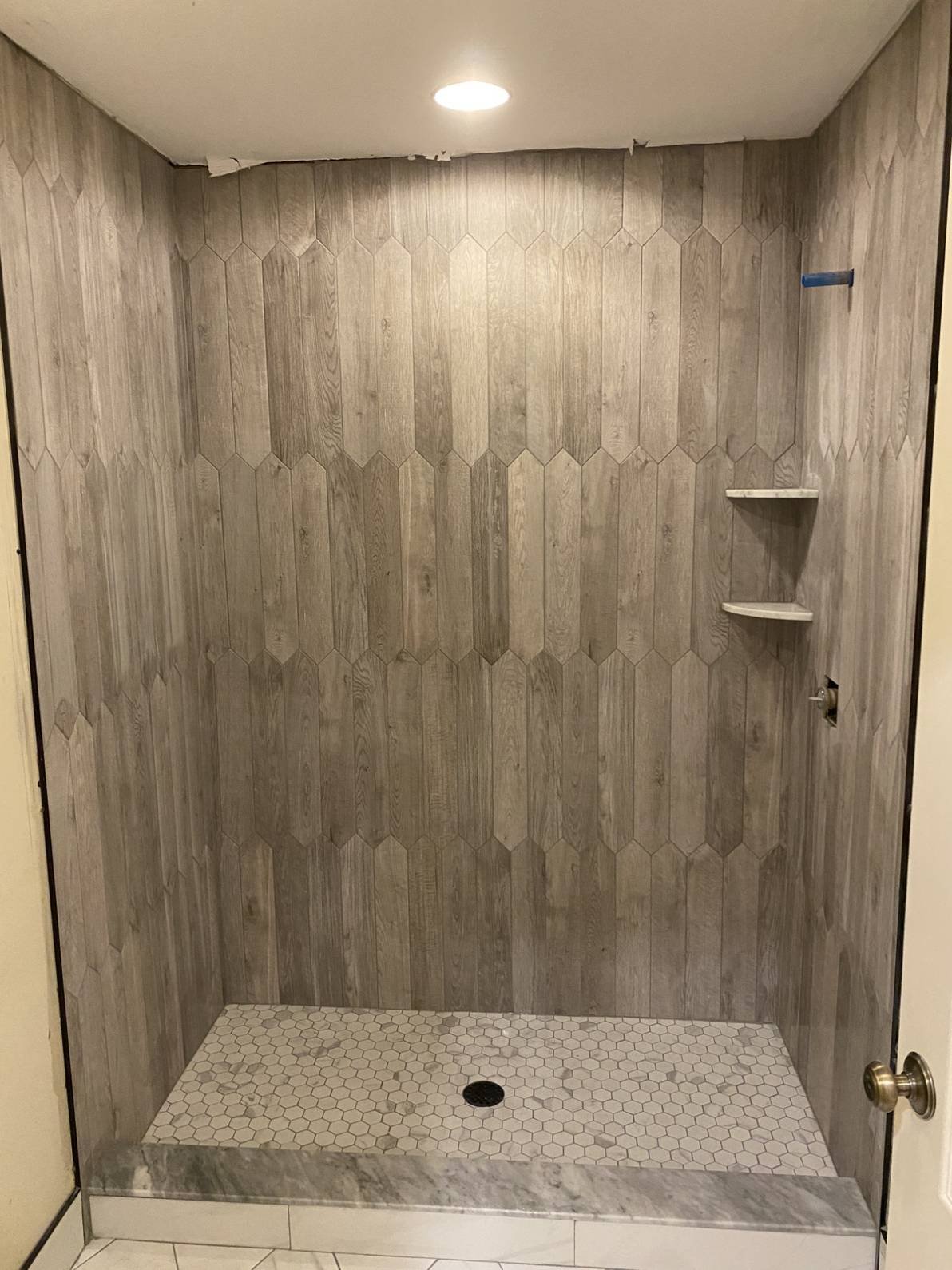 Empty walk-in shower with wood-look tile walls, small hexagon floor tiles, built-in shelves, and a ceiling light.