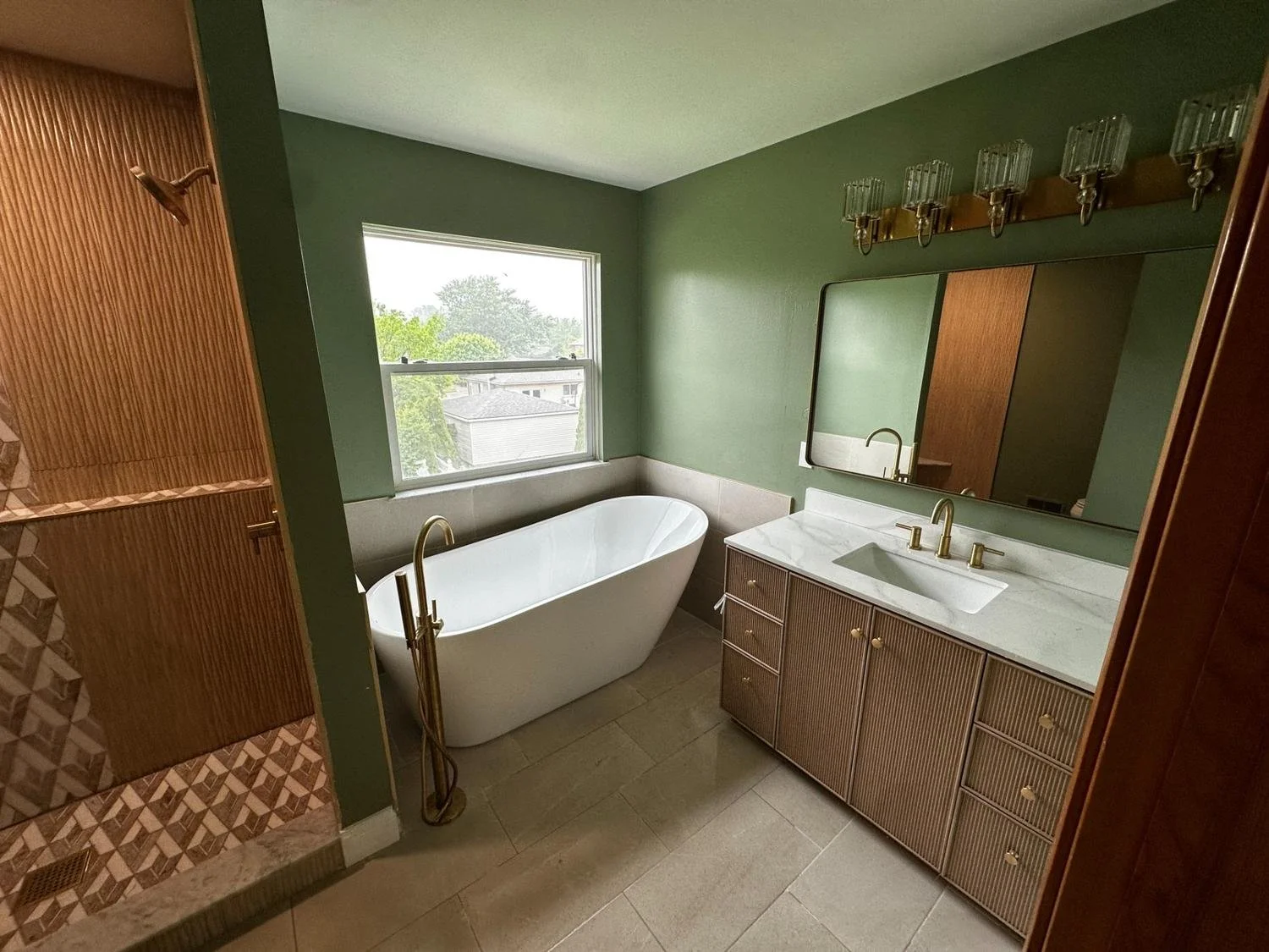 Modern bathroom with a white bathtub, a large wall mirror, green painted walls, a wooden vanity with a white marble countertop, and a window showing green trees outside.