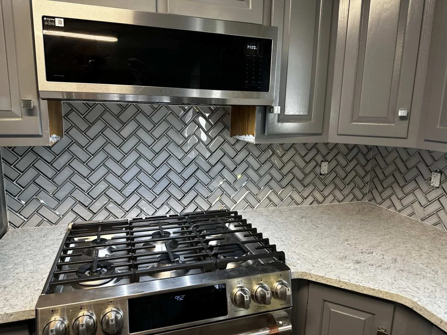 Kitchen stove with stainless steel and a granite countertop, grey kitchen cabinets, and a mosaic tile backsplash, with a microwave oven mounted above the stove.