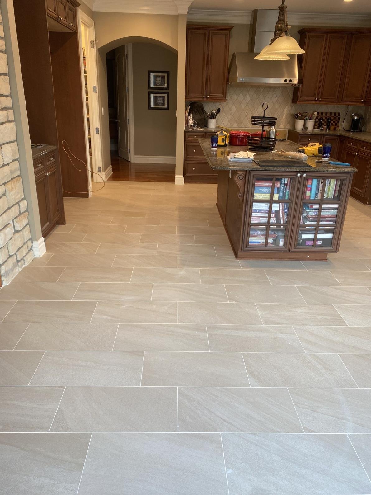 Empty kitchen with beige tile flooring, dark wood cabinets, granite countertops, a kitchen island with glass-front cabinets filled with books, and some tools and items on the counter. In the background, part of an arched doorway and a hallway with fr