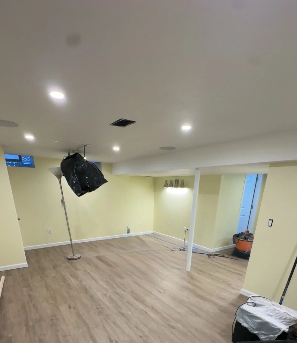 Undeveloped basement room with yellow walls, wood floors, and ceiling lights. Contains construction materials and equipment, including a lamp, a portable heater, and a black plastic bag hanging from the ceiling.