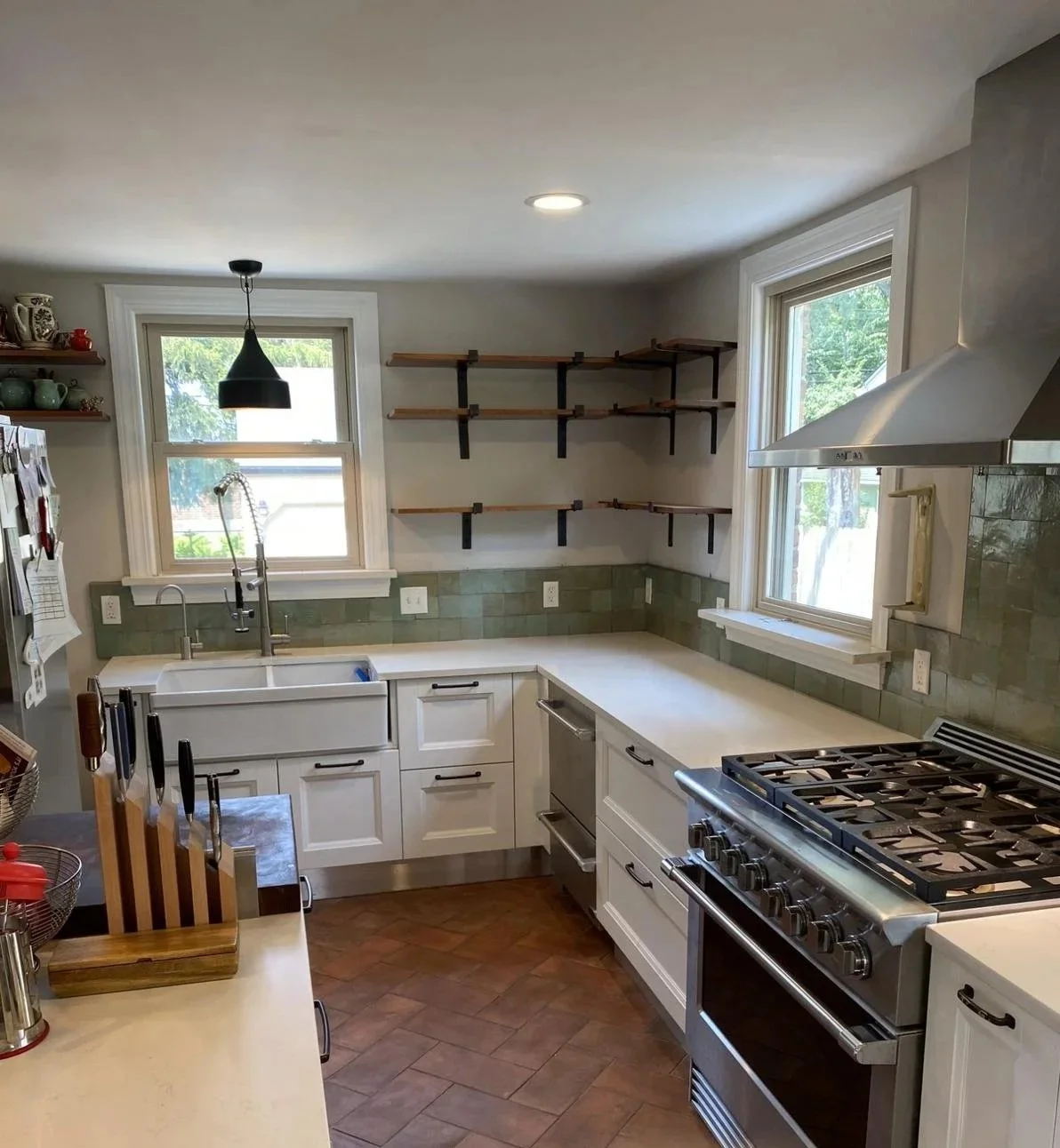 Empty kitchen with white cabinets, green tiled backsplash, stainless steel appliances, open shelves, and two windows letting in natural light.