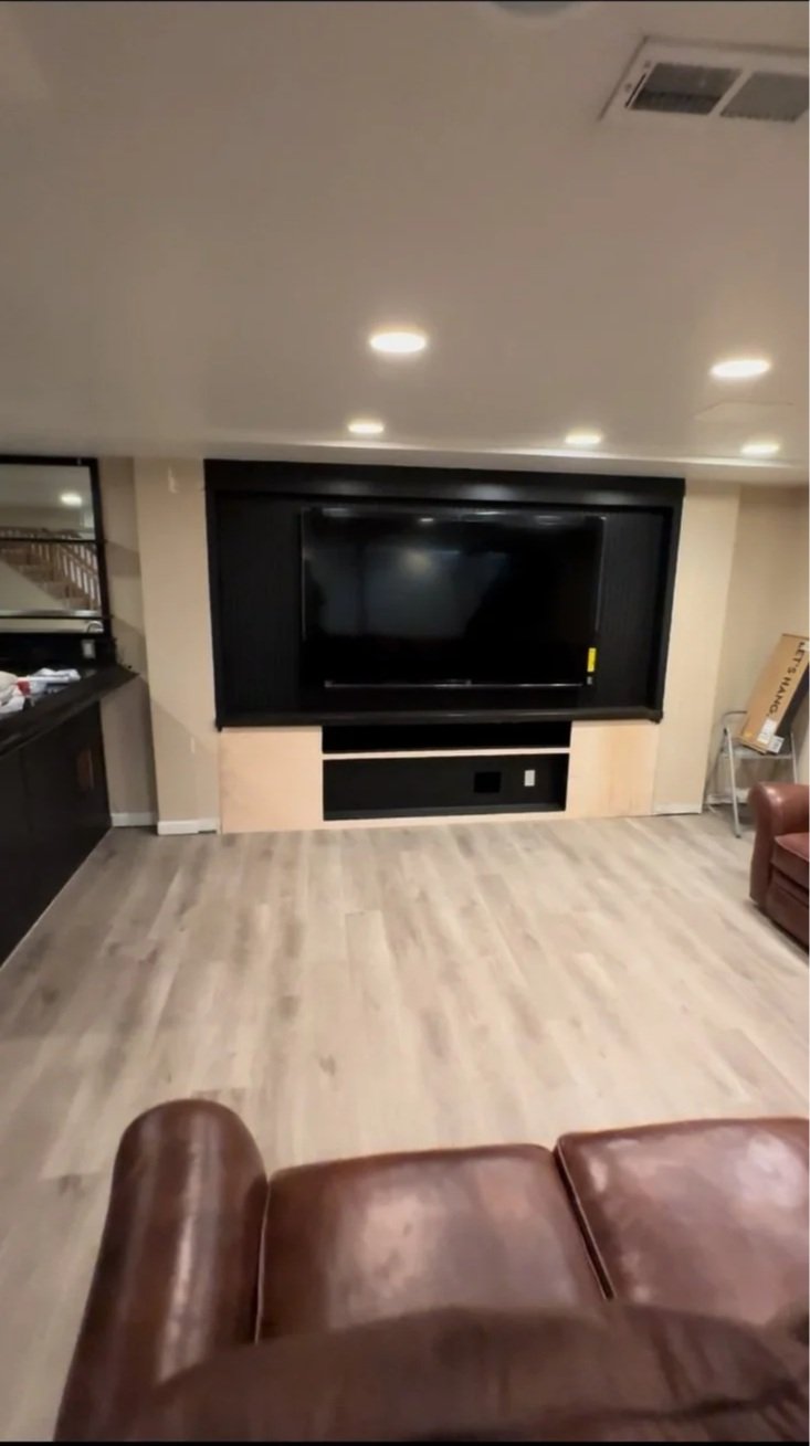 A living room with a large flat-screen TV mounted on the wall and a beige wooden panel beneath it, surrounded by a black frame. The room has light-colored hardwood flooring, a brown leather couch in the foreground, and a small side table or cabinet o