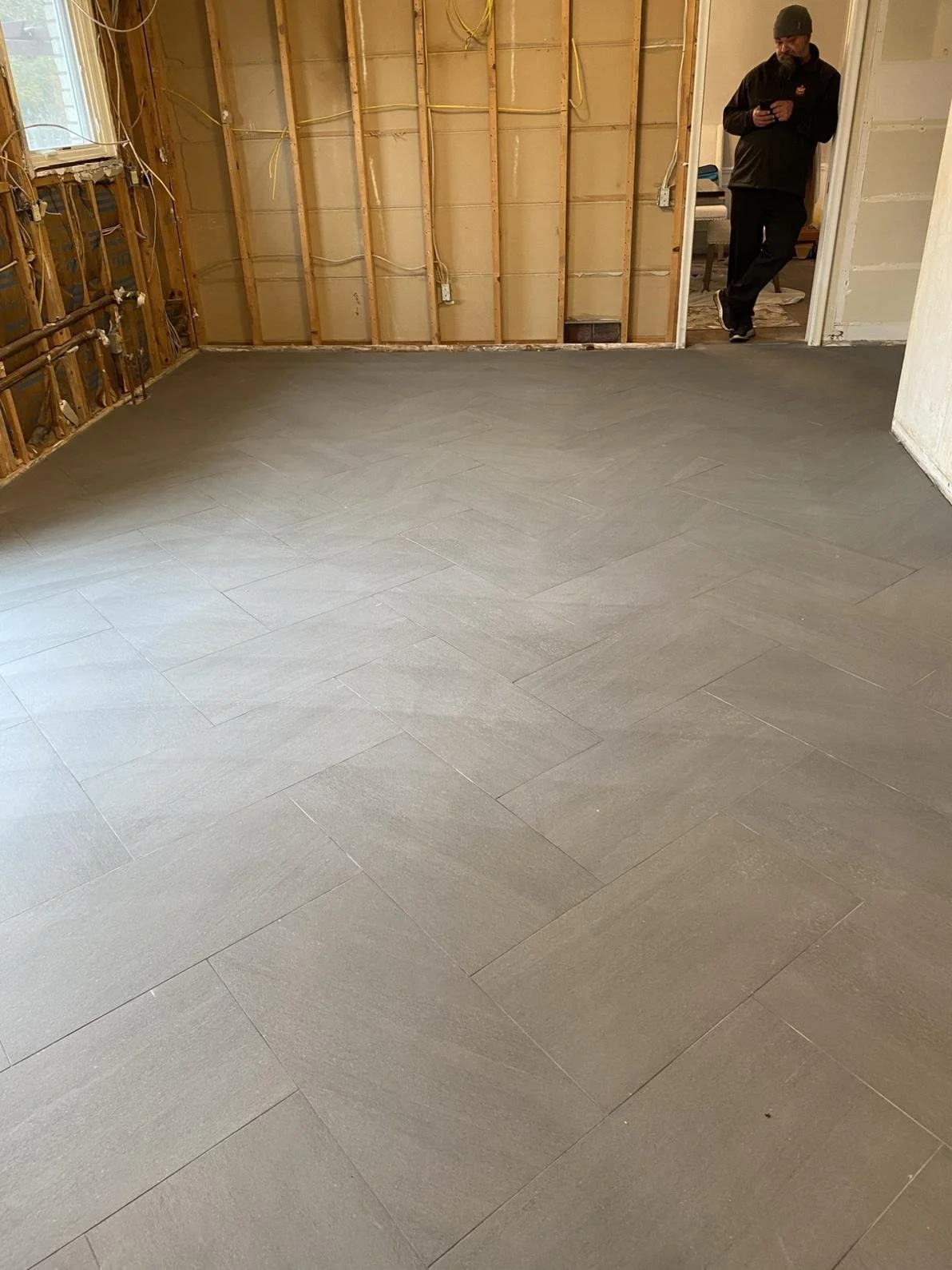 Interior of a room under renovation with a newly installed gray tile floor, exposed wall studs, and a person standing in a doorway looking at a phone.