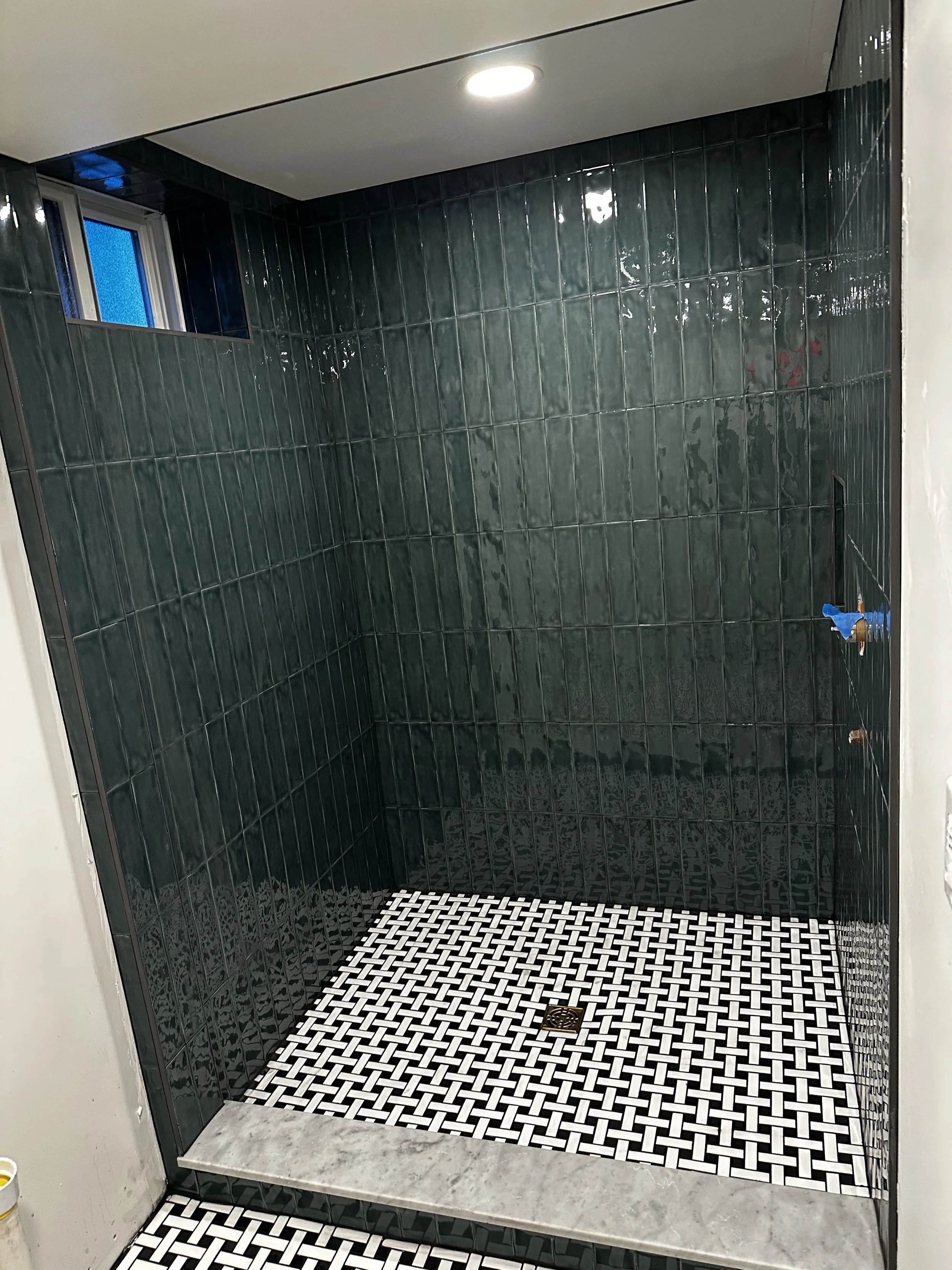 A newly tiled walk-in shower area with dark green glossy tiles on the walls, white and black patterned tiles on the floor, a small window to the left, and a ceiling light.
