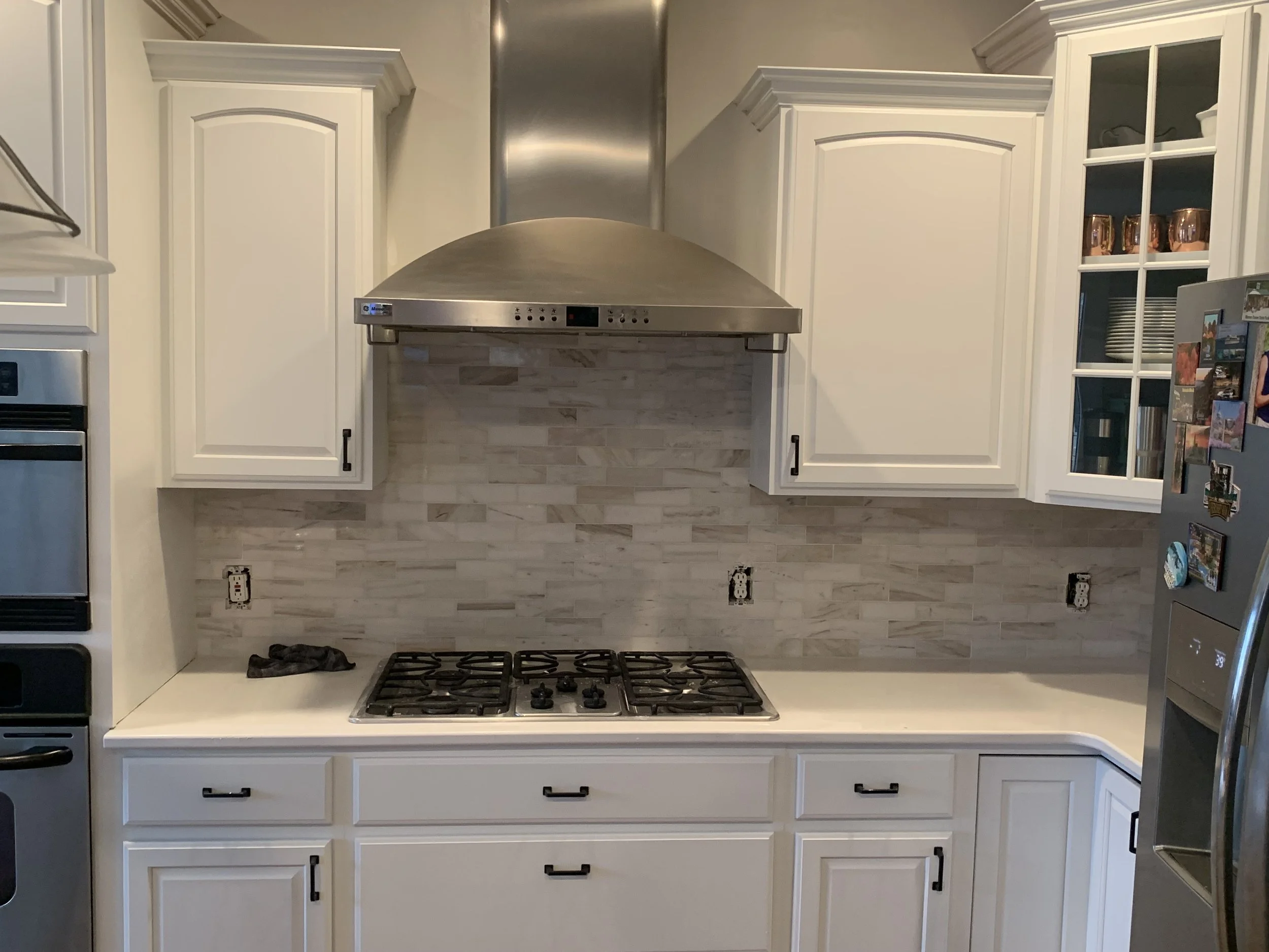 Kitchen with white cabinets, gas stove, stainless steel vent hood, and a light-colored tiled backsplash.
