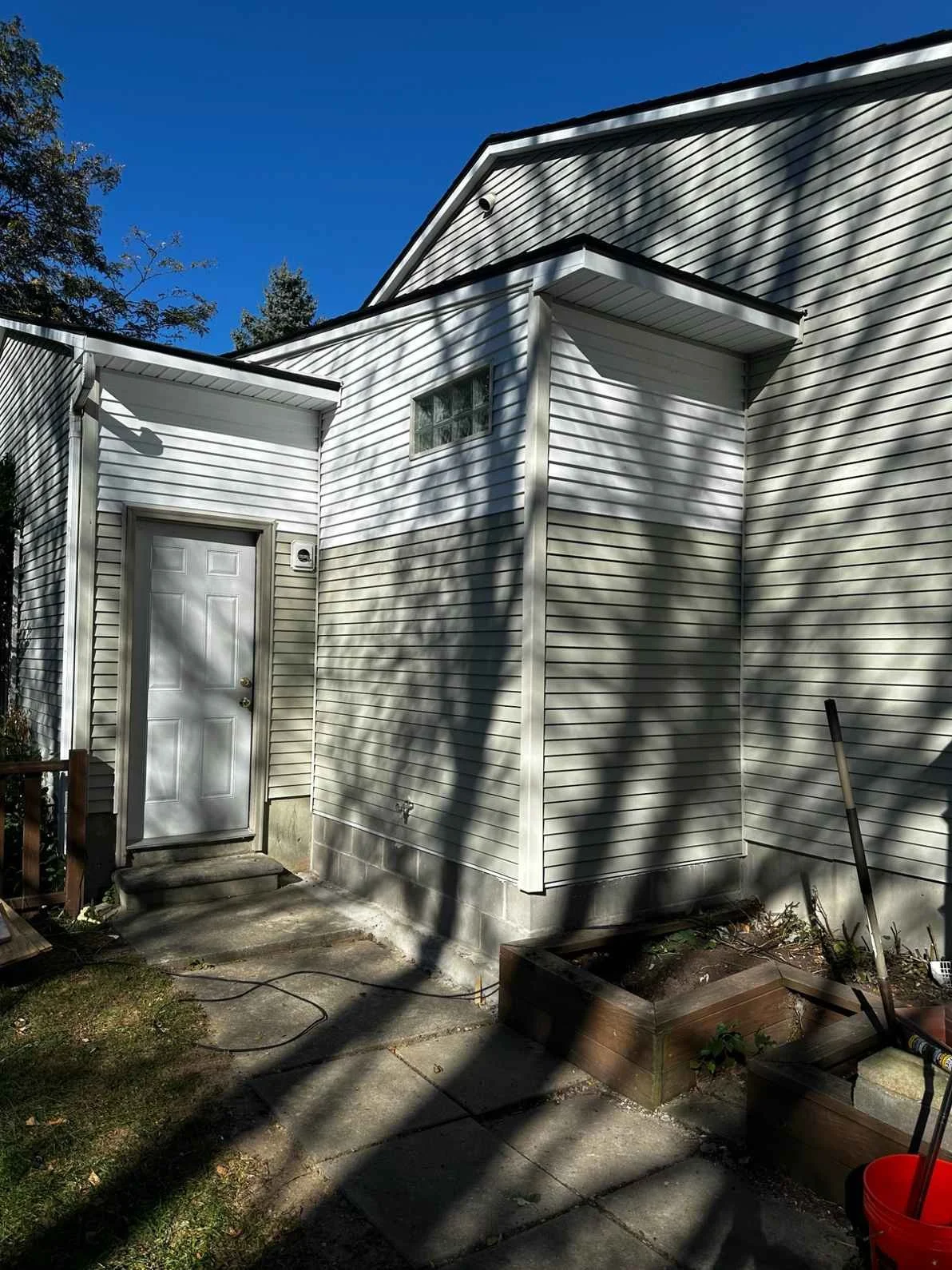 Side view of a house with beige vinyl siding, a white door with a small set of concrete steps, and a small window above it. Shadows of trees are cast on the house. There is a garden bed and some gardening tools on the ground.