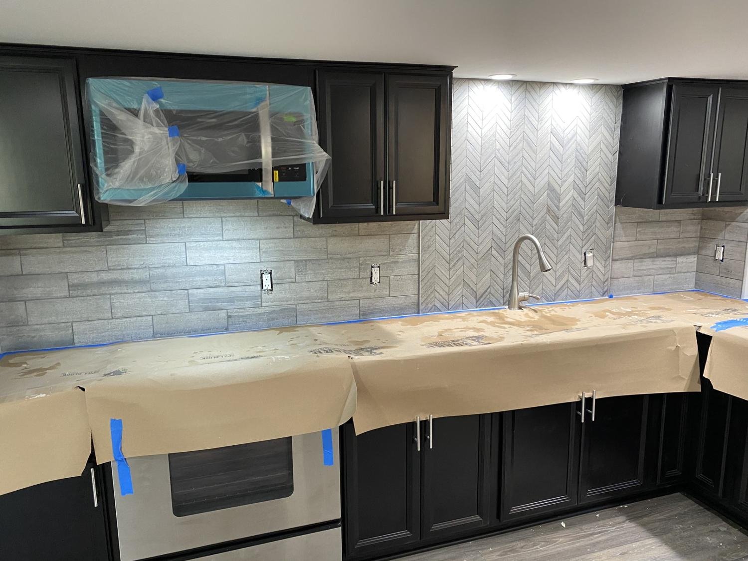 Kitchen under renovation with black cabinets, gray backsplash tiles, and protective paper on the counters, with some wiring and a range hood wrapped in plastic.