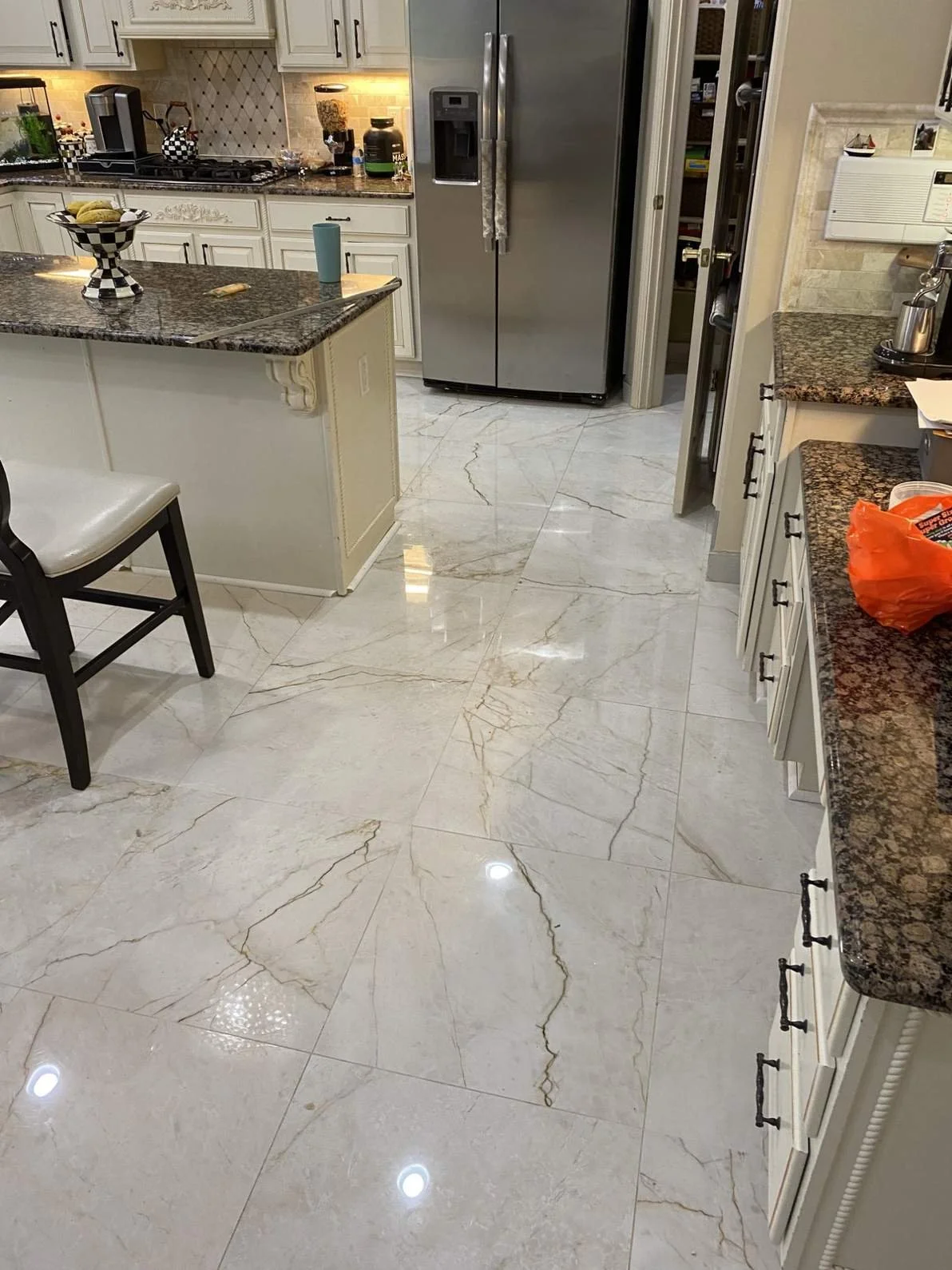 Kitchen with white cabinets and granite countertops, stainless steel refrigerator, tiled marble floor, and some groceries in an orange plastic bag on the right counter.