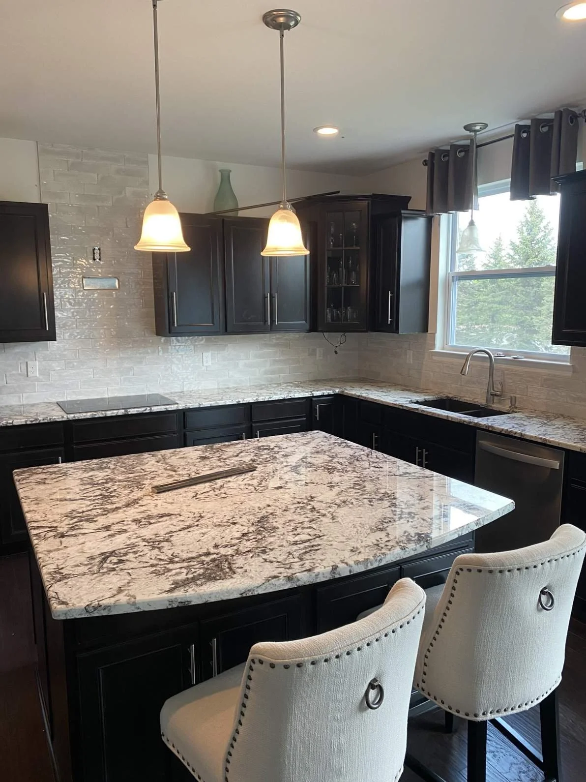 Modern kitchen with dark cabinets, a large marble island, and two white upholstered chairs.