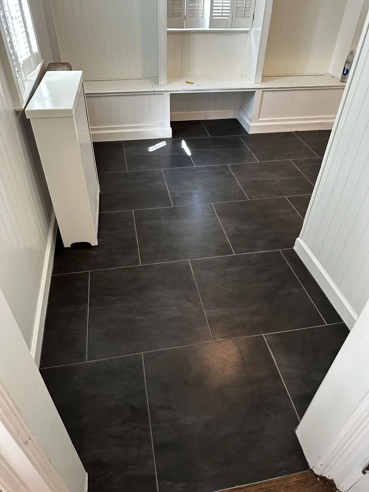 A room with large dark gray floor tiles, white paneled walls, a white radiator cover on the left, and a window with white blinds at the back.