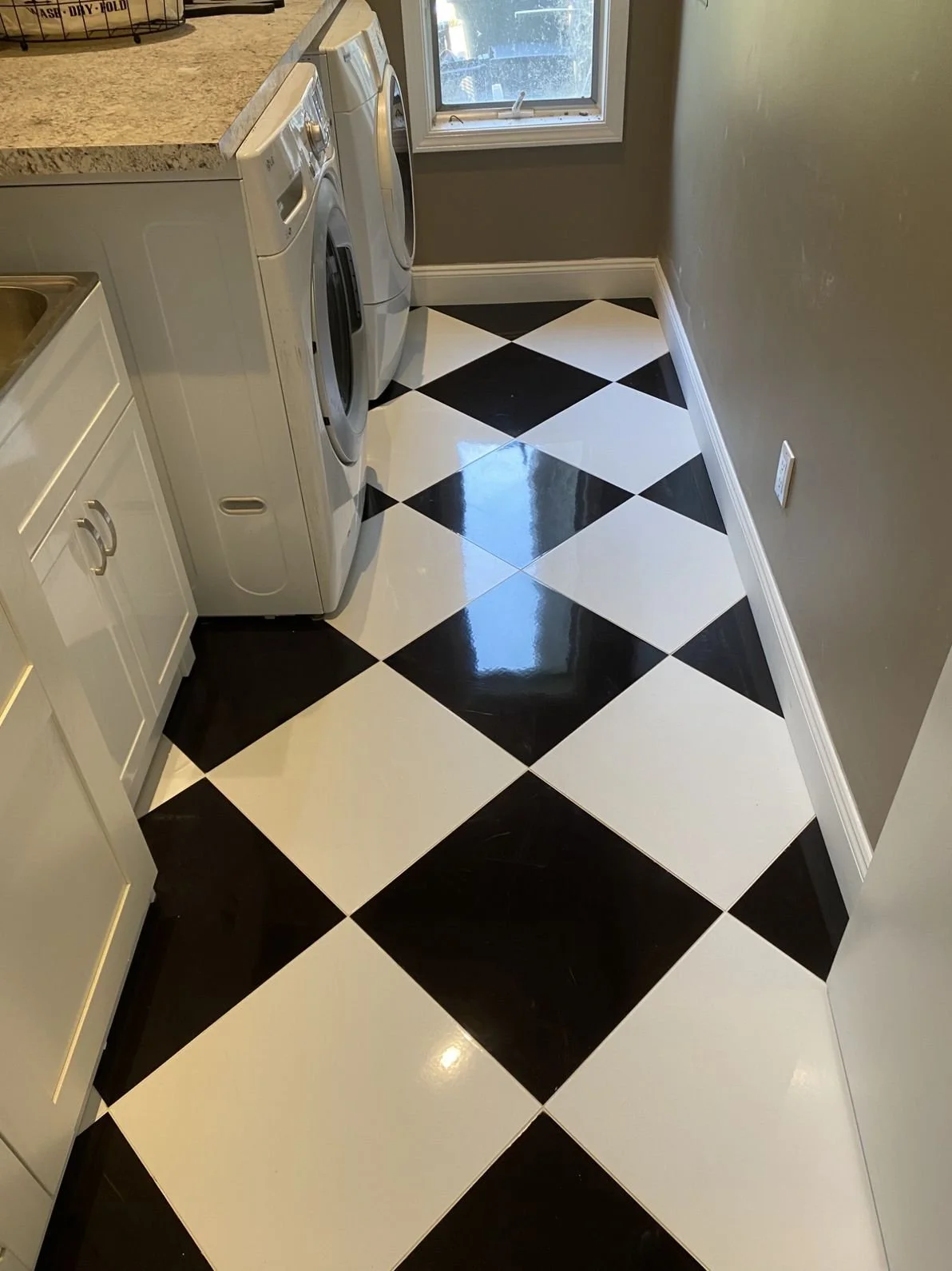 A laundry room with black and white checkered tile flooring, a white washer and dryer, part of a granite countertop, a small window, and a gray wall.