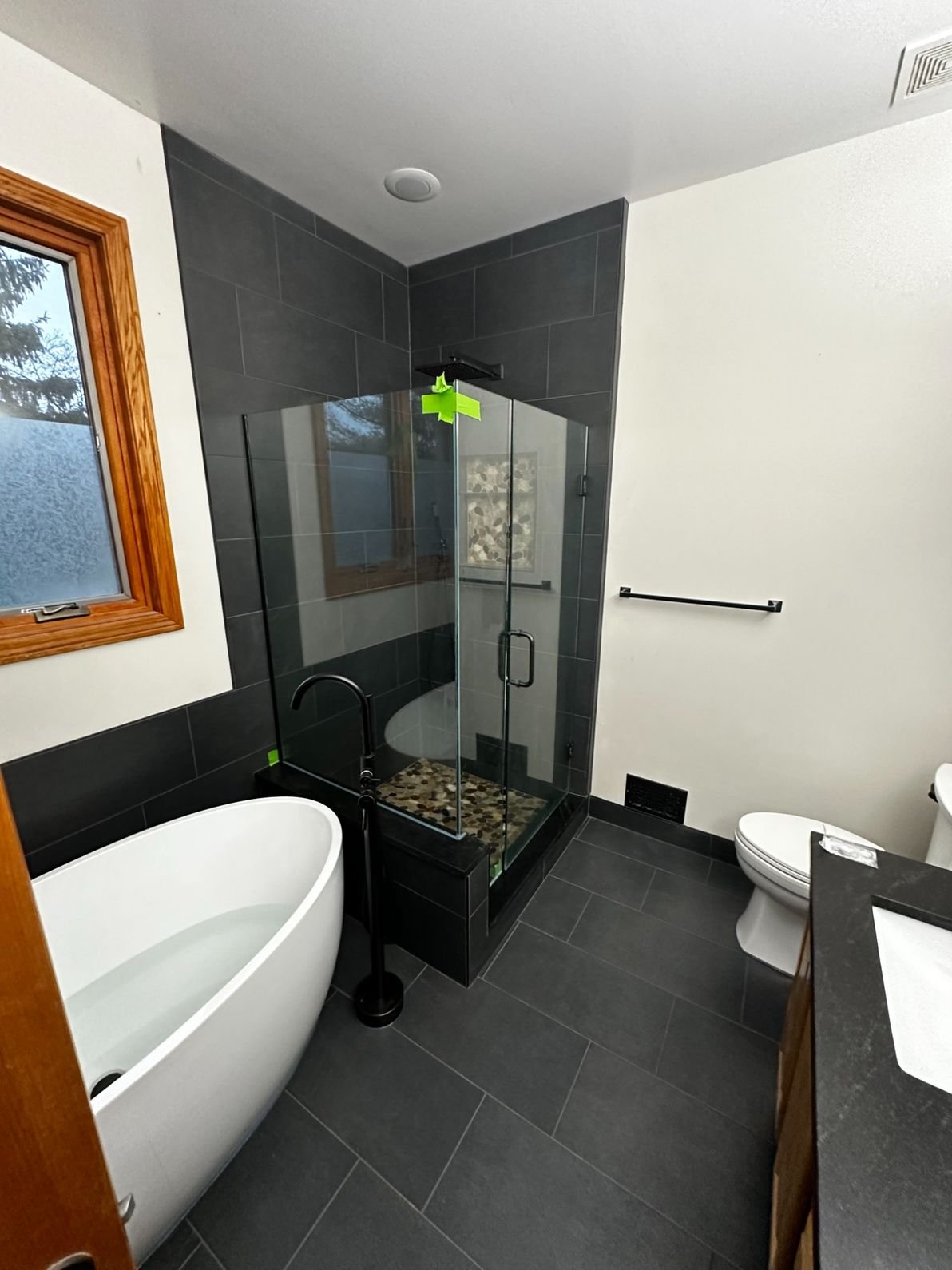 Modern bathroom with a freestanding bathtub, a glass-enclosed shower with black fixtures, a toilet, and a wooden-framed window. Black tile flooring and wall accents.