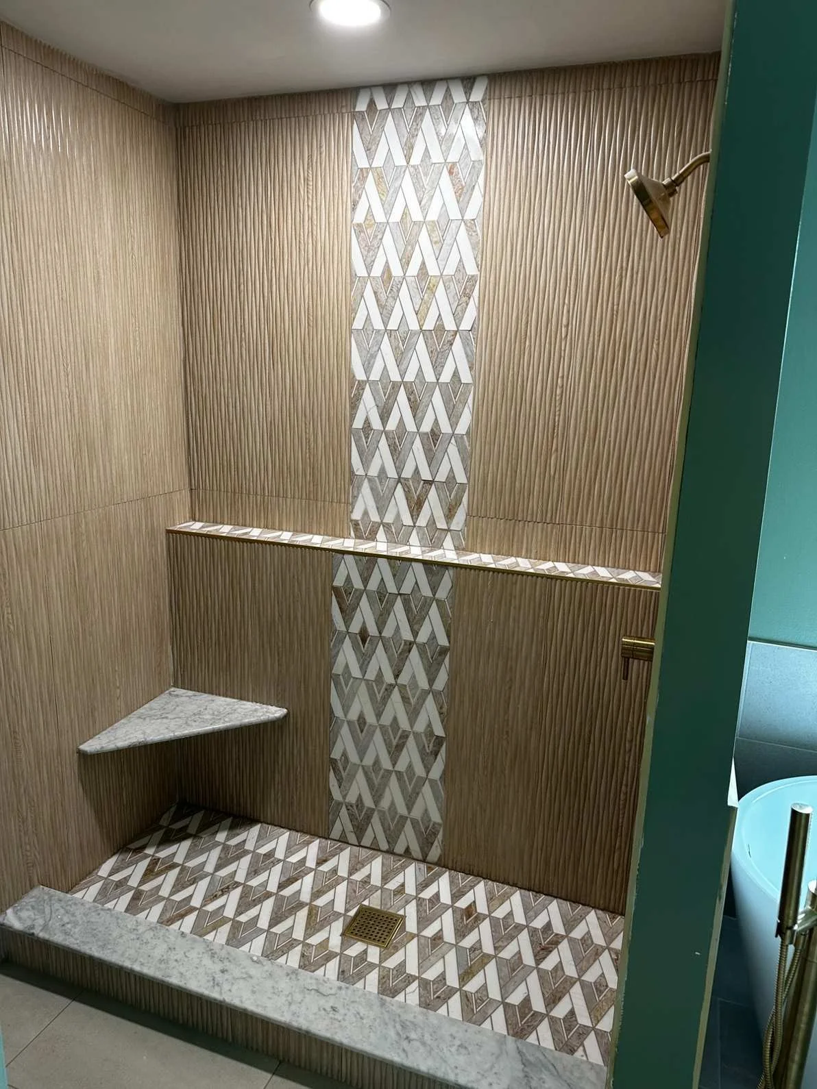 A modern shower area with distinctive wall and floor tiles, a marble shelf in the corner, and a brass shower head. The wall features a vertical strip of geometric tiles embedded in textured wood-patterned wall panels.