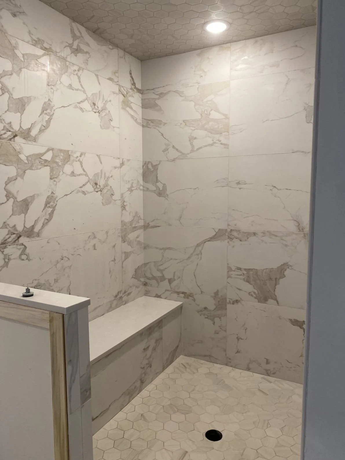 A shower area with marble-like tiles on the walls and small hexagonal tiles on the floor, with a built-in bench and a ceiling light.