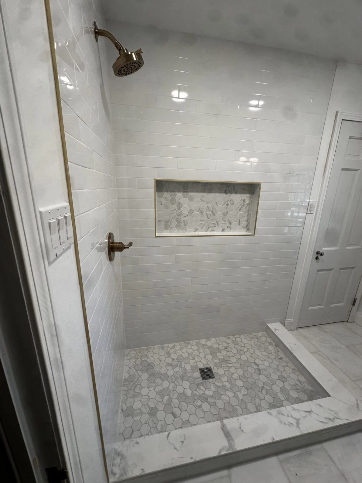 A modern shower with white subway tiles, a built-in niche with hexagonal tiles, and hexagonal floor tiles in shades of gray, framed by white marble slab.
