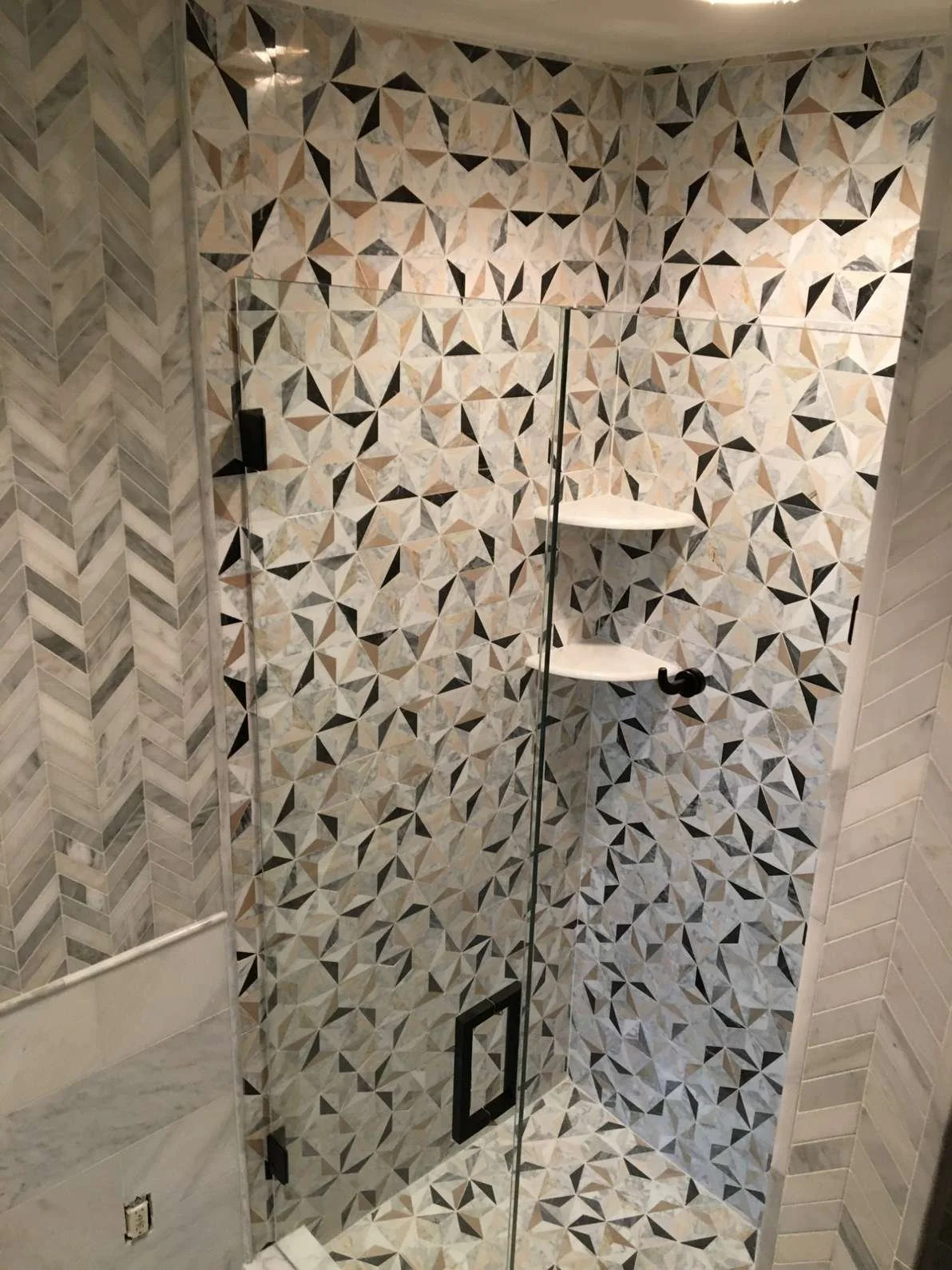A modern shower with glass door and geometric tile pattern on the walls and floor. There are two white corner shelves and a black door handle.