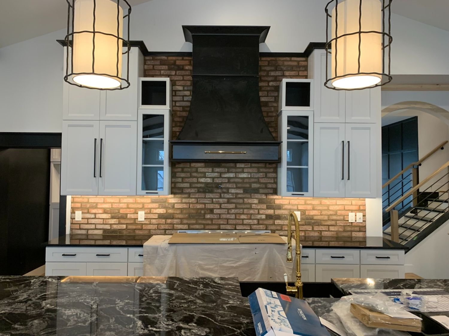 Kitchen with white cabinets, black countertops, a brick backsplash, and a black range hood. Pendant lights hang from the ceiling, and a gold faucet is visible over a sink. There are stairs on the right side.