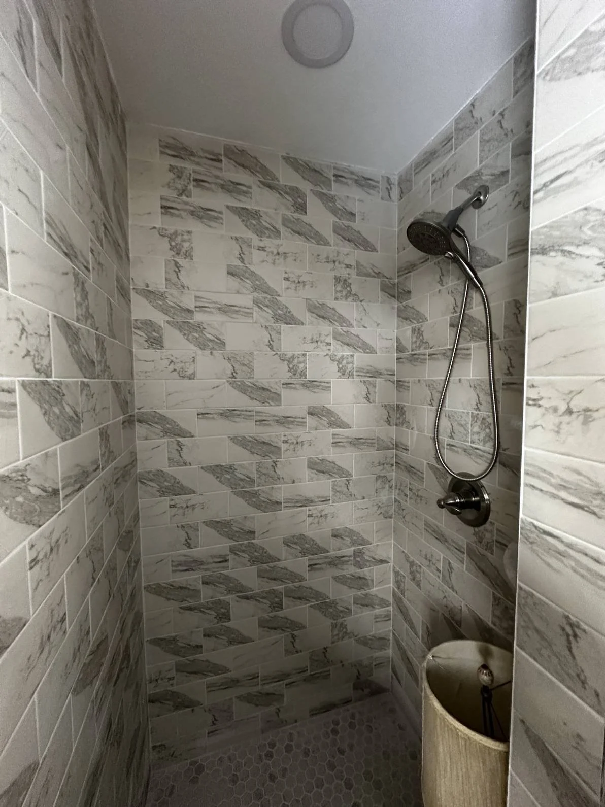 Shower area with marble-patterned tiles on the walls, a showerhead, and a small round container below.
