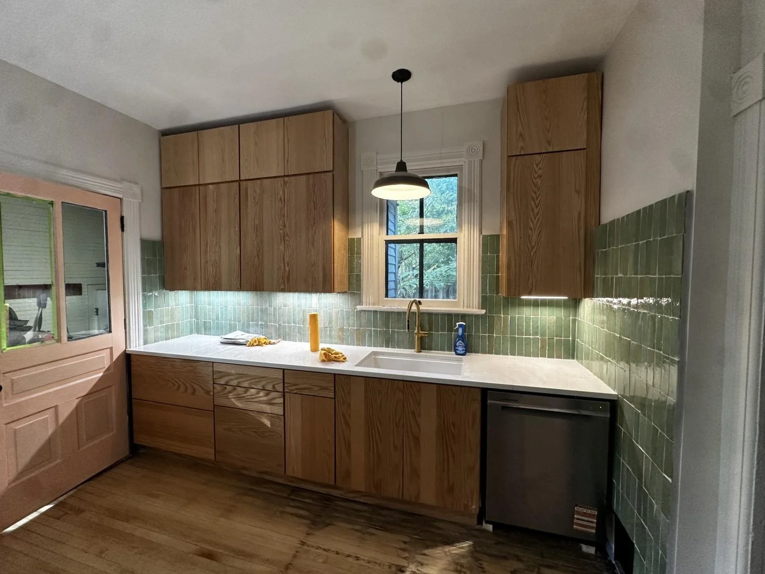 Kitchen with wood cabinets, green tiled backsplash, white countertop, window above sink, and wooden floor. Cleaning supplies on countertop.