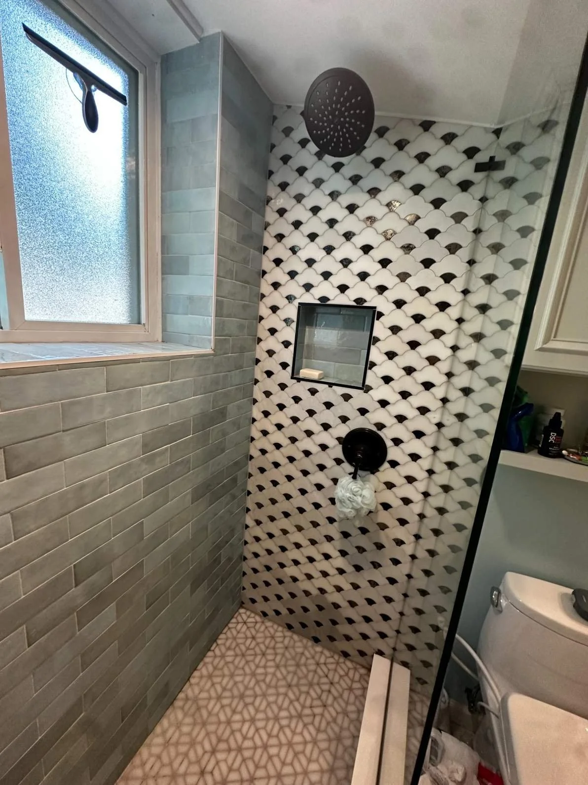 Bathroom shower area with distinctive patterned black and white tile on the wall, a black showerhead, a built-in shelf with soap, and a frosted window.