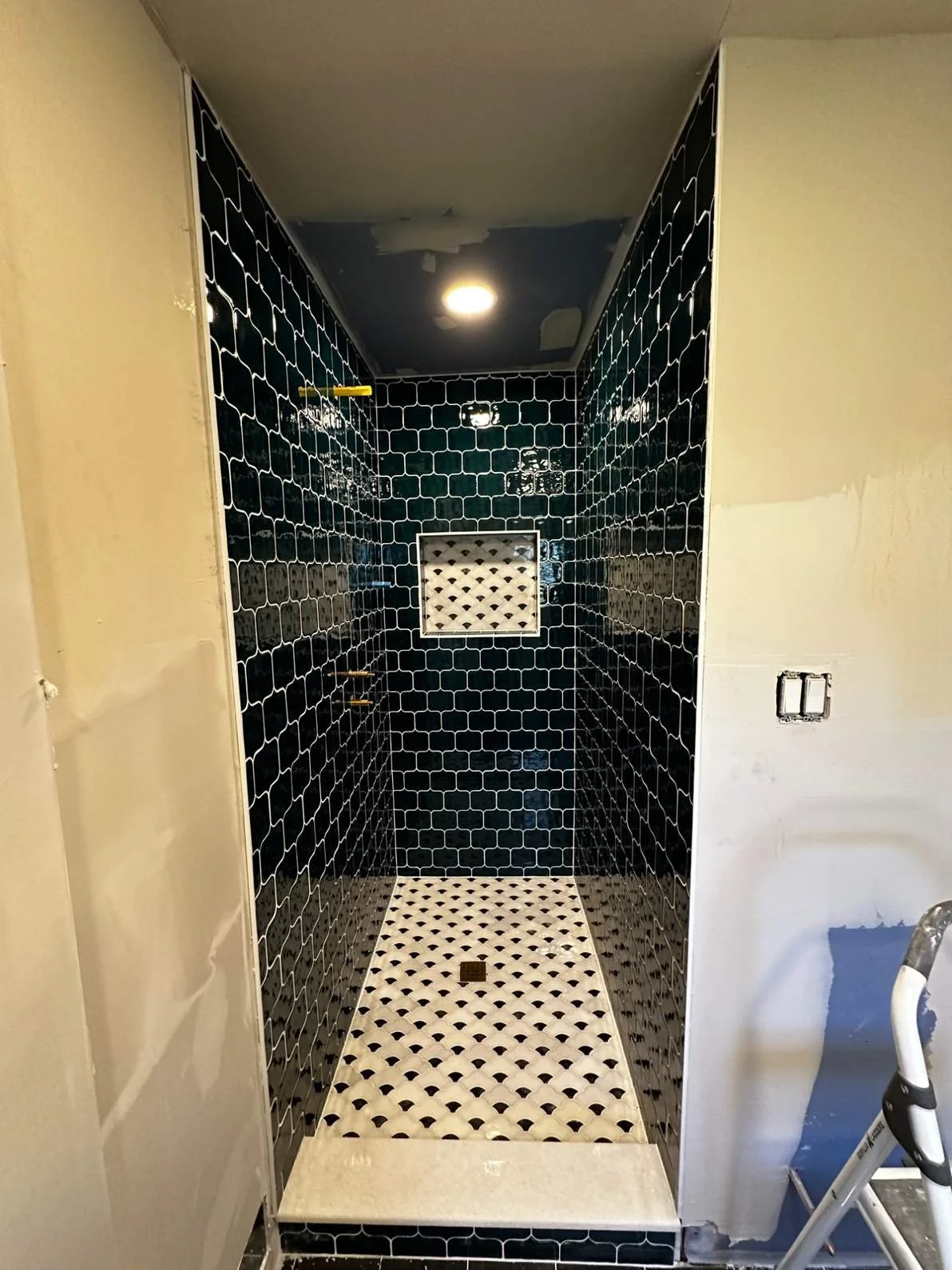 A small shower area with black glossy tiled walls and a white and black patterned floor. There is a small window on the back wall and a ceiling light fixture above.