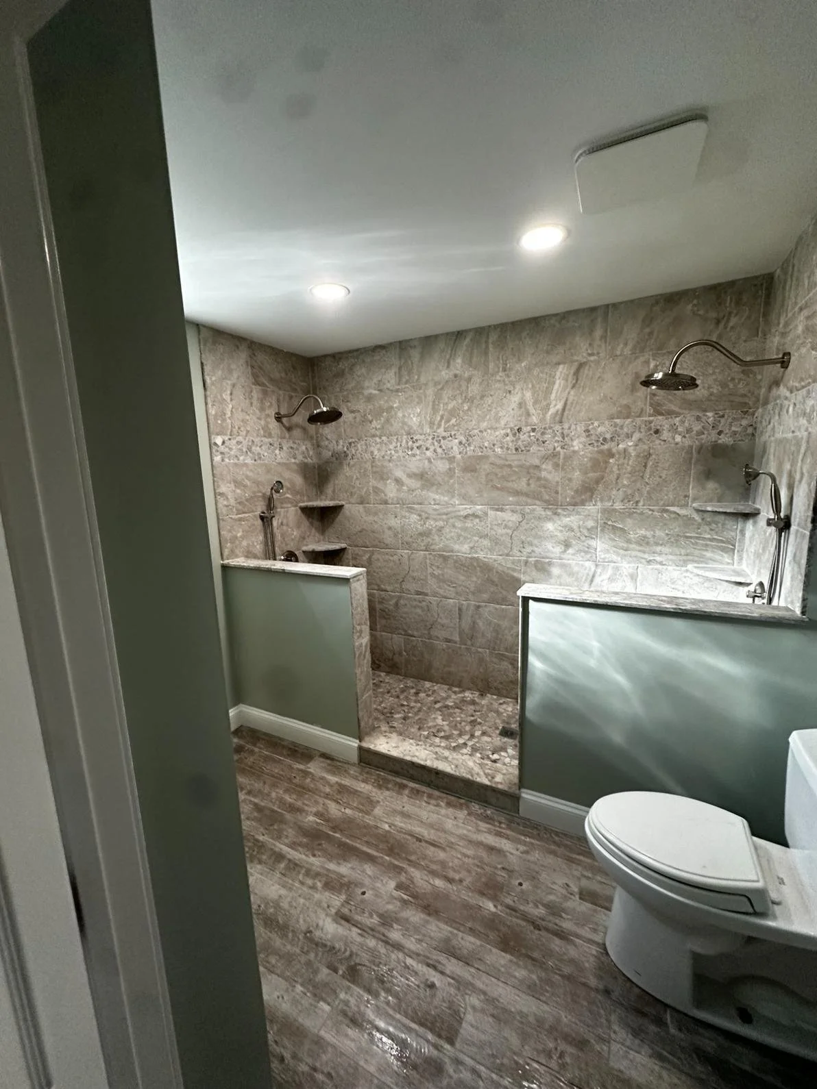 Bathroom with two showers, each with a showerhead on the wall, a small toilet, and wood-look flooring.