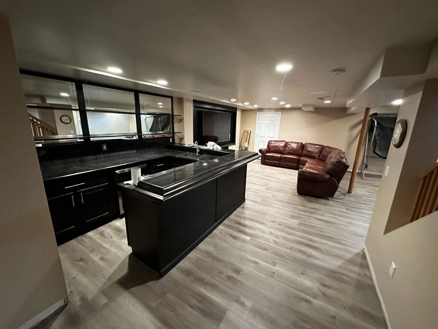 Empty basement with black kitchen island, brown leather sectional sofa, wood-look flooring, and minimal decor.