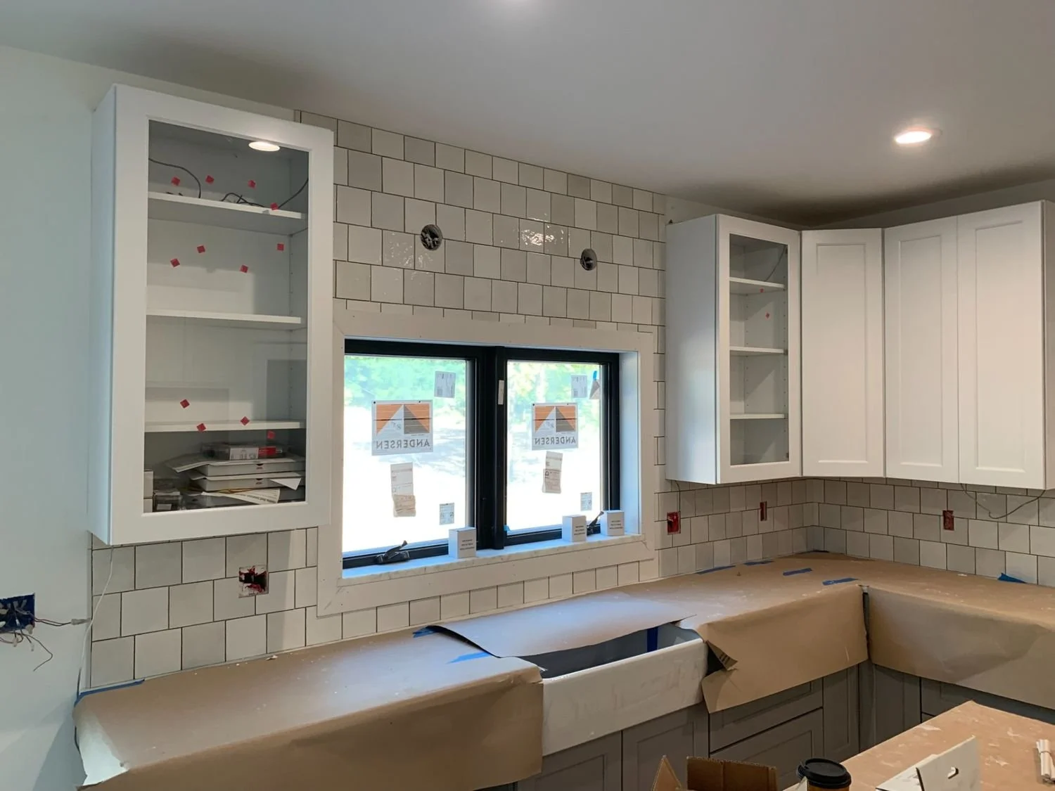 Kitchen under construction with white cabinetry, upper cabinets with glass doors, a window, and beige tiled walls.