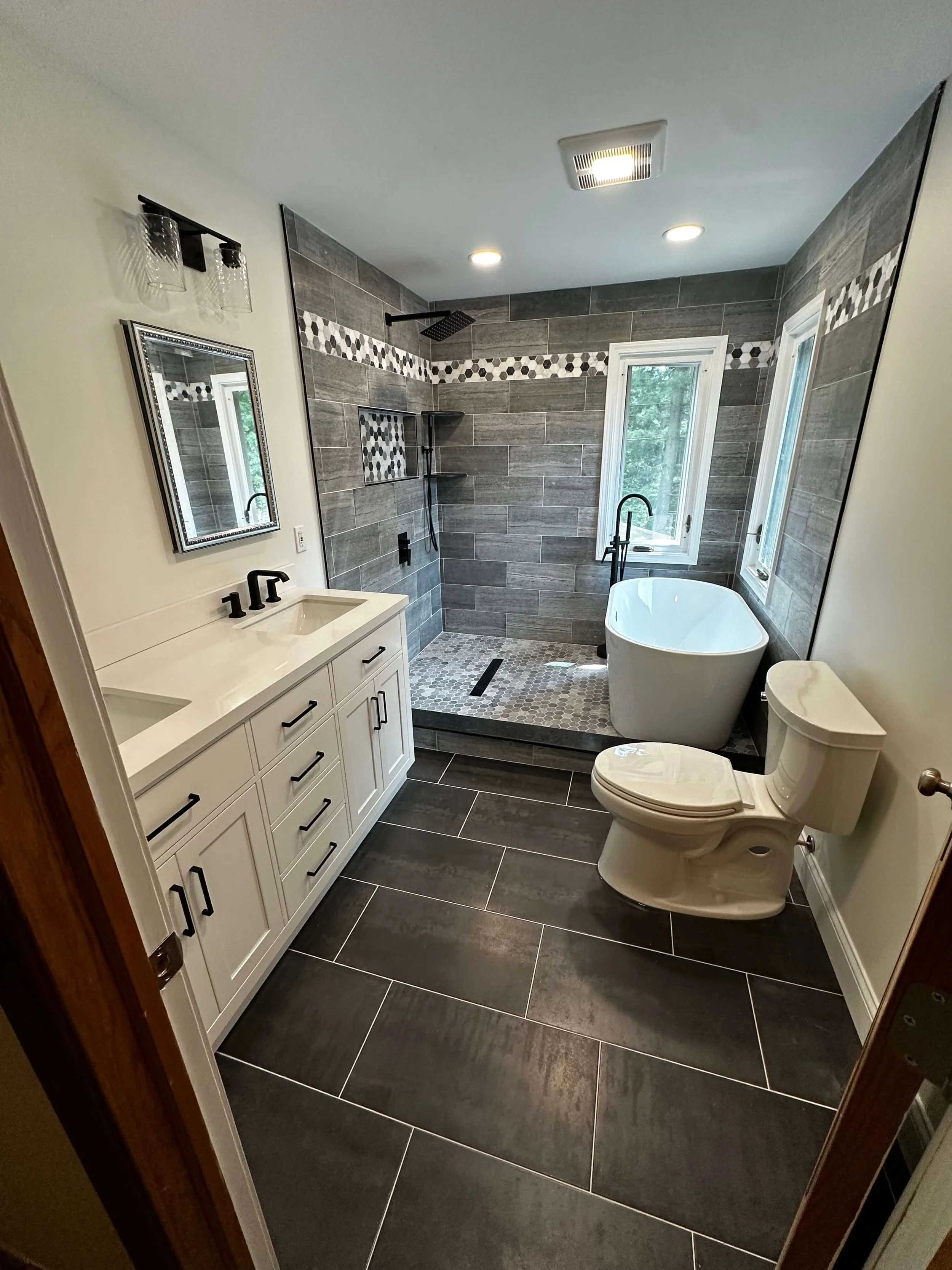 Modern bathroom with black and white fixtures, a vanity with a double sink, a walk-in shower with gray tile walls, a freestanding bathtub, and two windows.