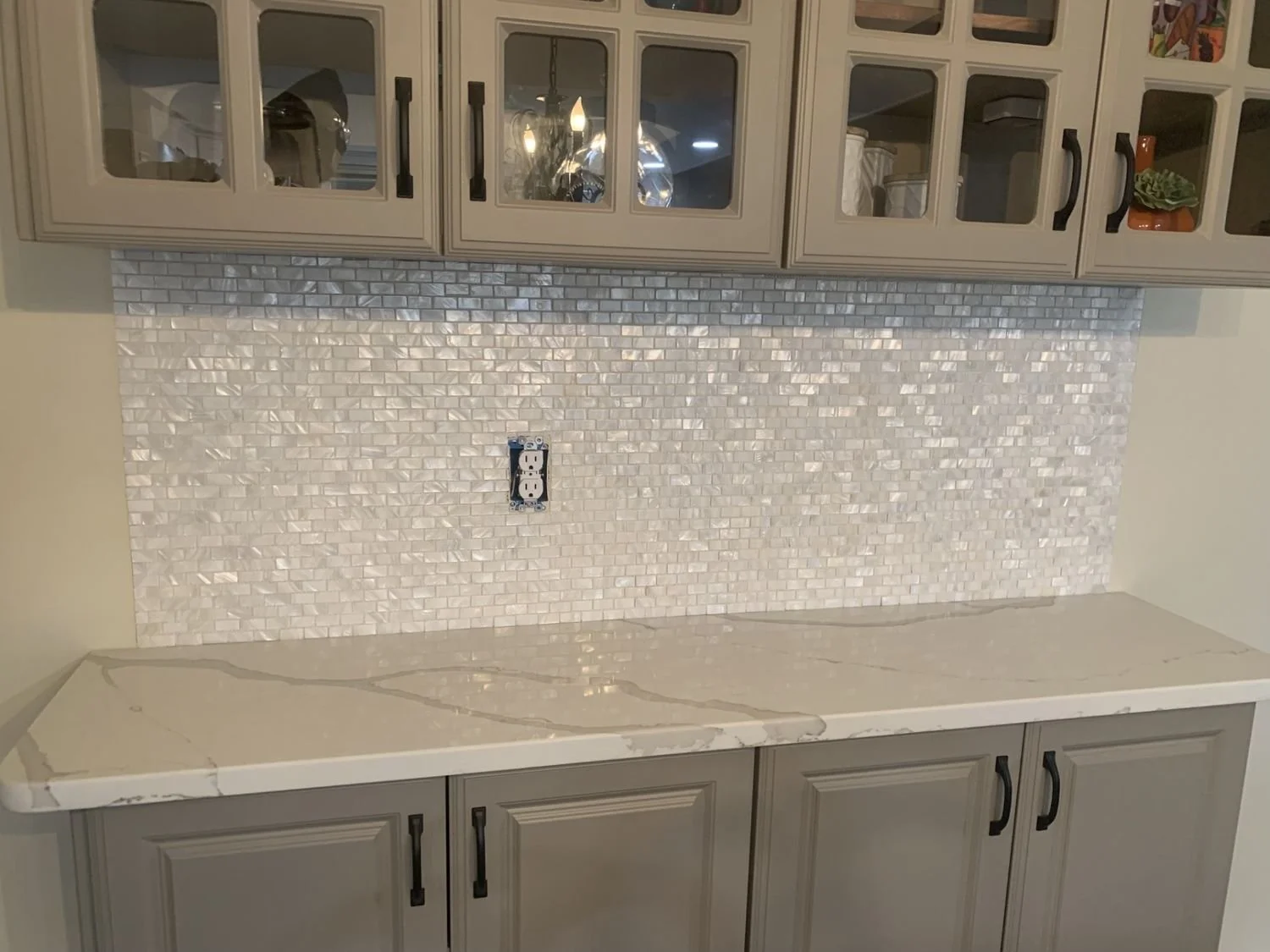 Kitchen counter with a white marble surface, beige cabinets with black handles, a tiled backsplash made of small iridescent white tiles, and an electrical outlet in the wall behind the counter.