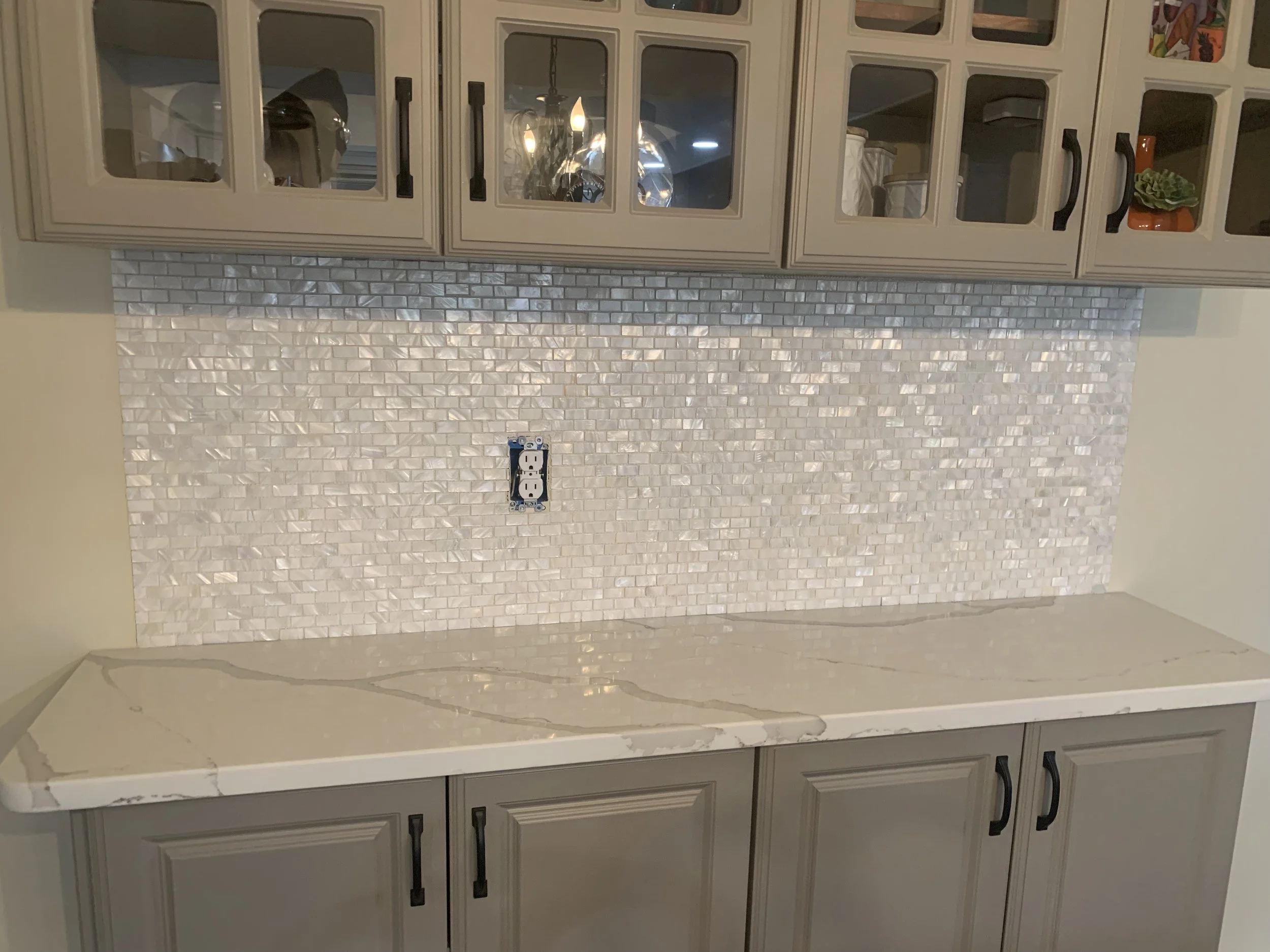 Kitchen backsplash with white mosaic tile behind a white marble countertop and beige cabinets with black handles.