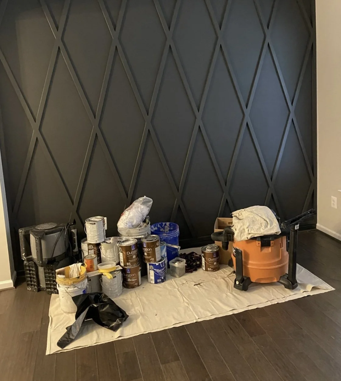 A wall with a geometric diamond pattern painted in dark color. Various painting supplies including cans of paint, a paint sprayer, and a small paint roller on a sheet of paper on the floor.