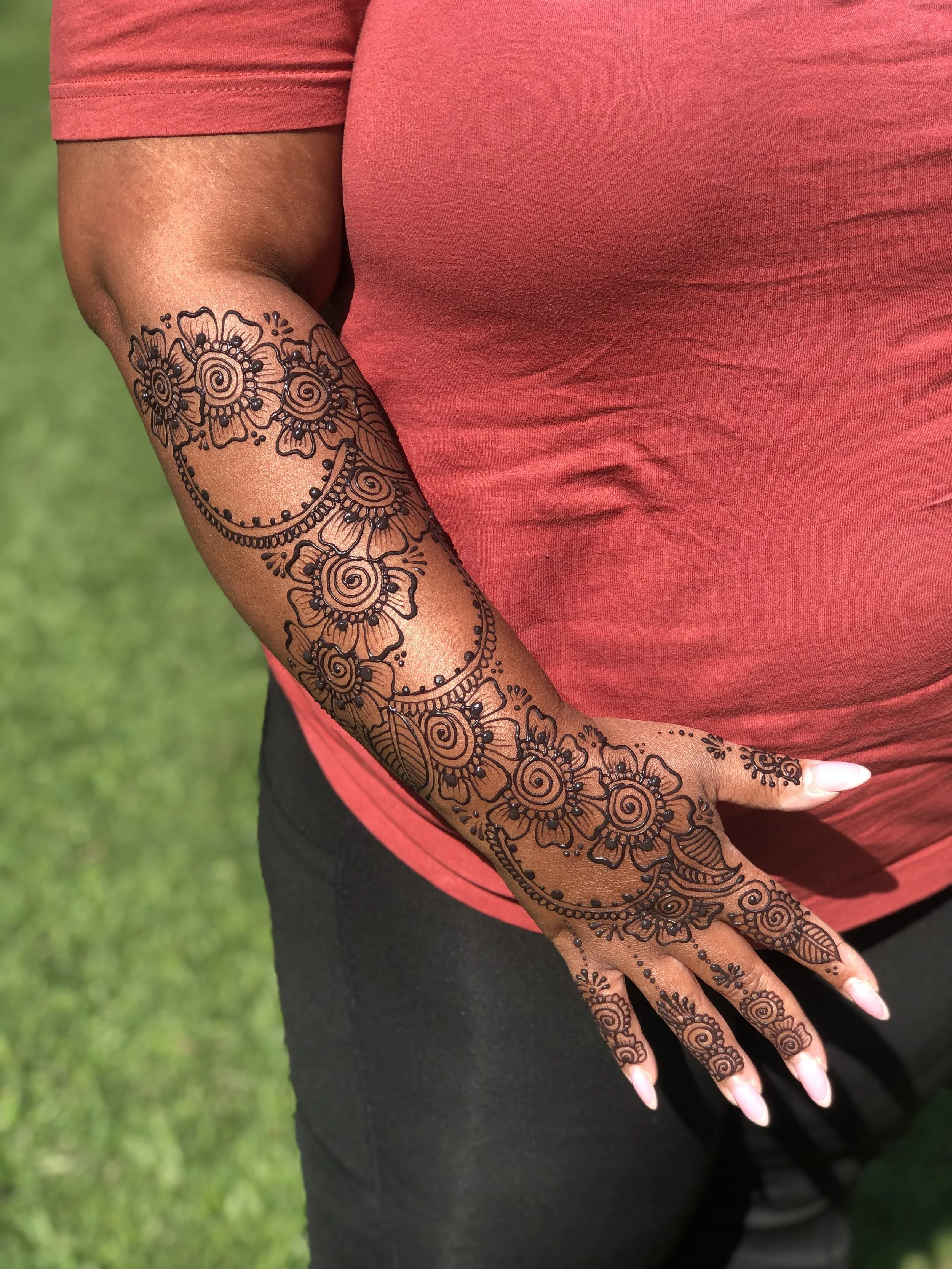 Close-up of a person with henna designs on their hand and forearm, wearing a red T-shirt and black pants, standing on green grass.