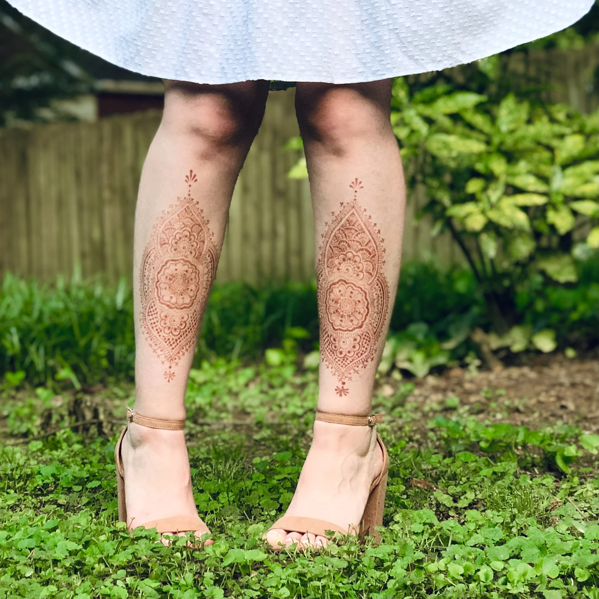 Close-up of the lower part of a person wearing beige sandals, patterned henna designs on legs, standing on green grass with a wooden fence and green foliage in the background.